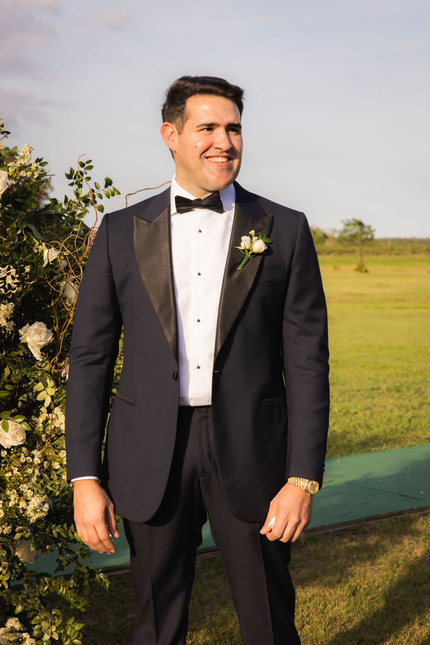 Groom in classic black tuxedo standing outdoors at sunset with boutonniere.