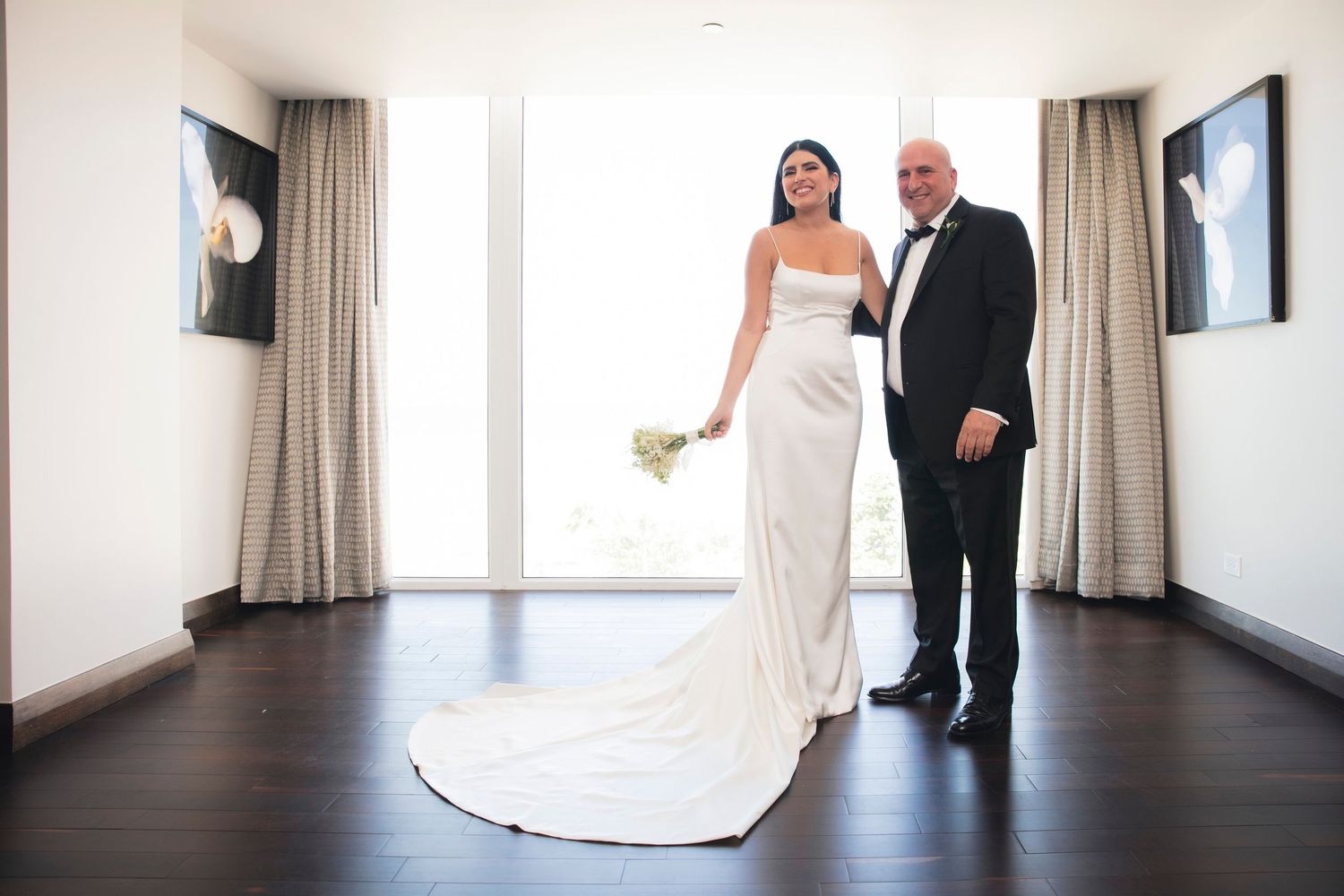 A couple in formal wedding attire stands in an elegant hallway with dark wood floors and cream curtains.