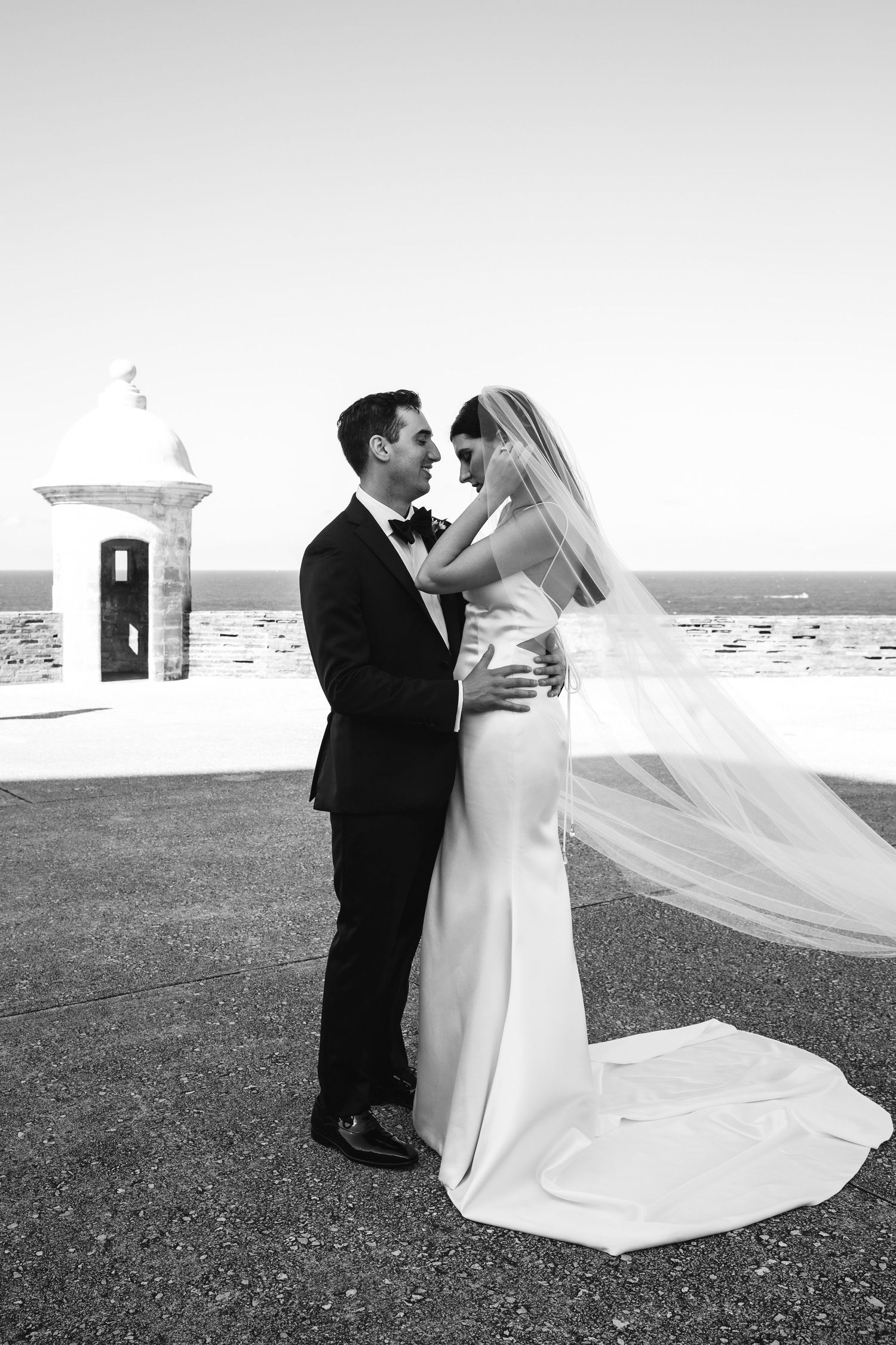 A series of black and white seaside wedding photos showing a couple embracing with a flowing veil in the breeze.