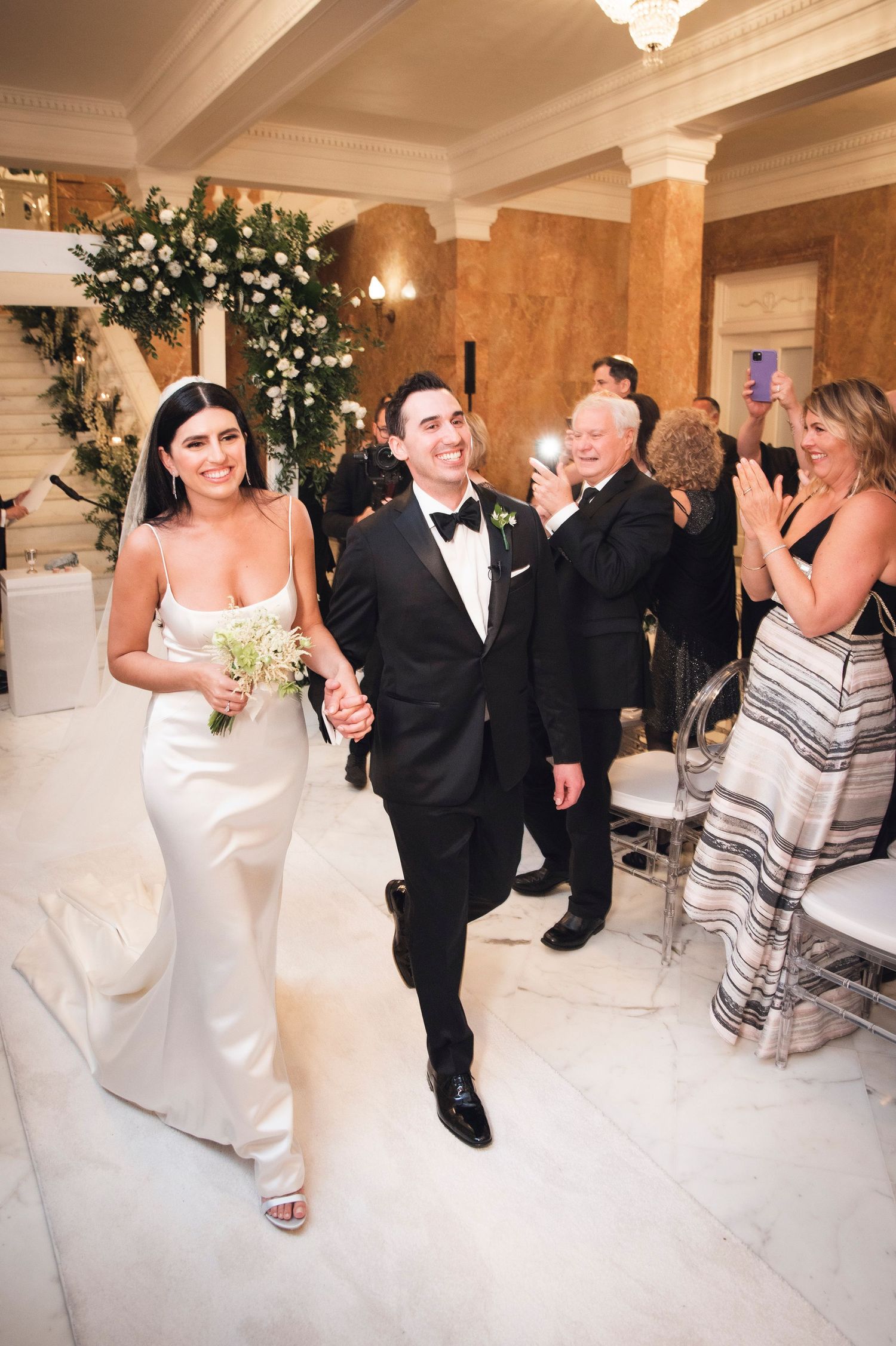 Joyful wedding dance sequence in an ornate white ballroom with guests looking on.