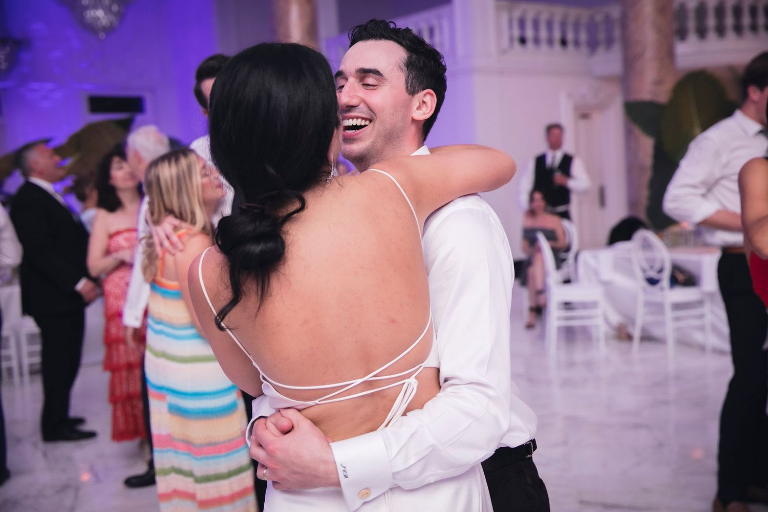 Wedding celebration with people dancing under purple lighting in an ornate ballroom setting.