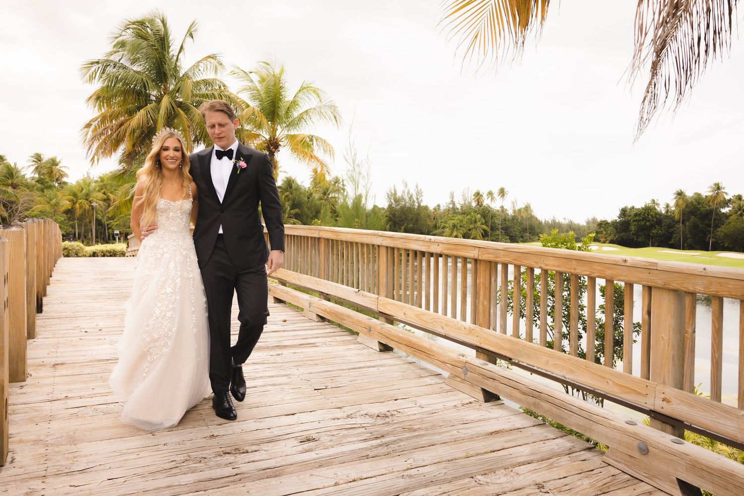 A couple walks along a wooden boardwalk surrounded by tropical palm trees at a beach wedding venue on a sunny day.