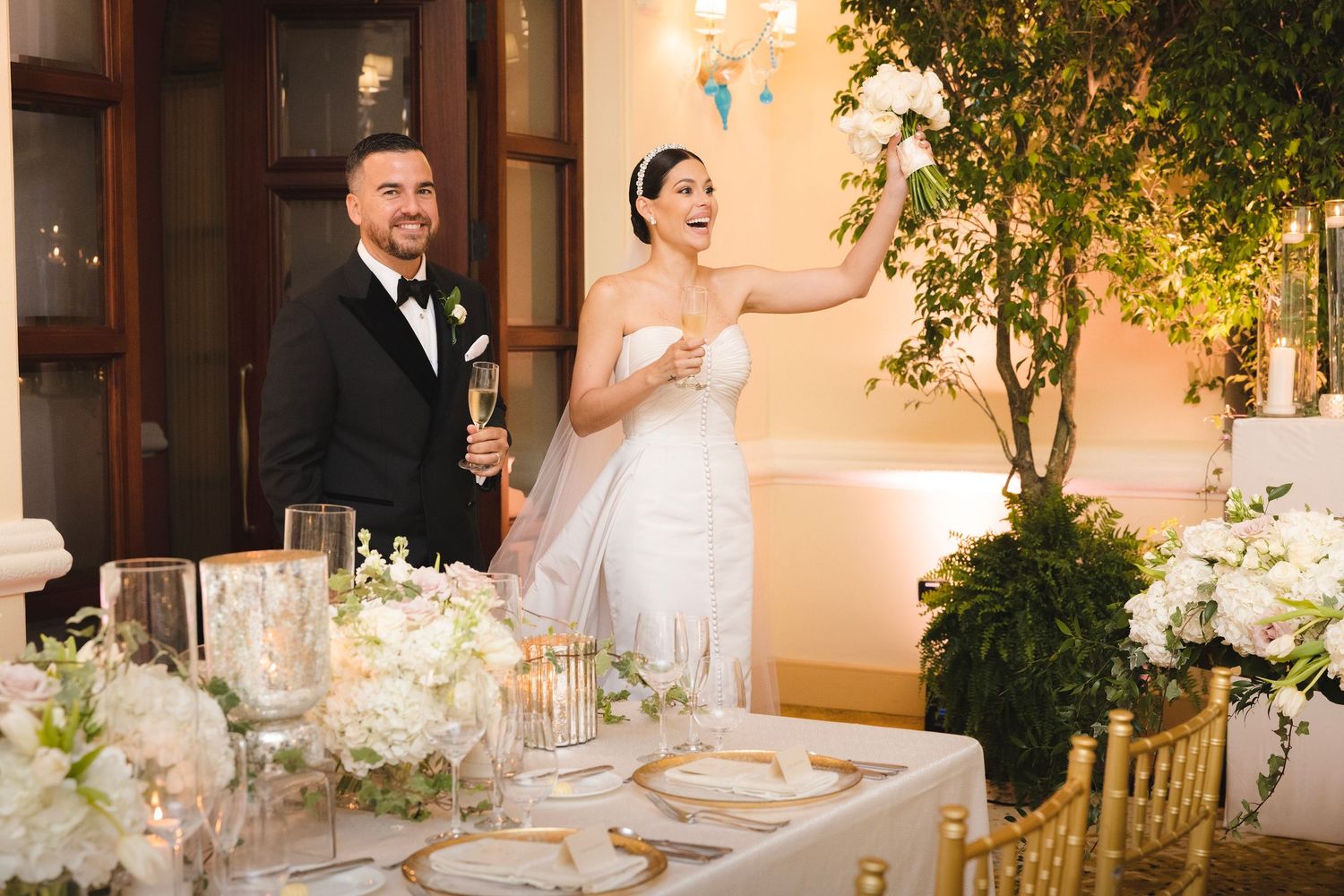 Wedding reception table set with white linens and floral centerpieces in an elegant indoor venue at dusk.