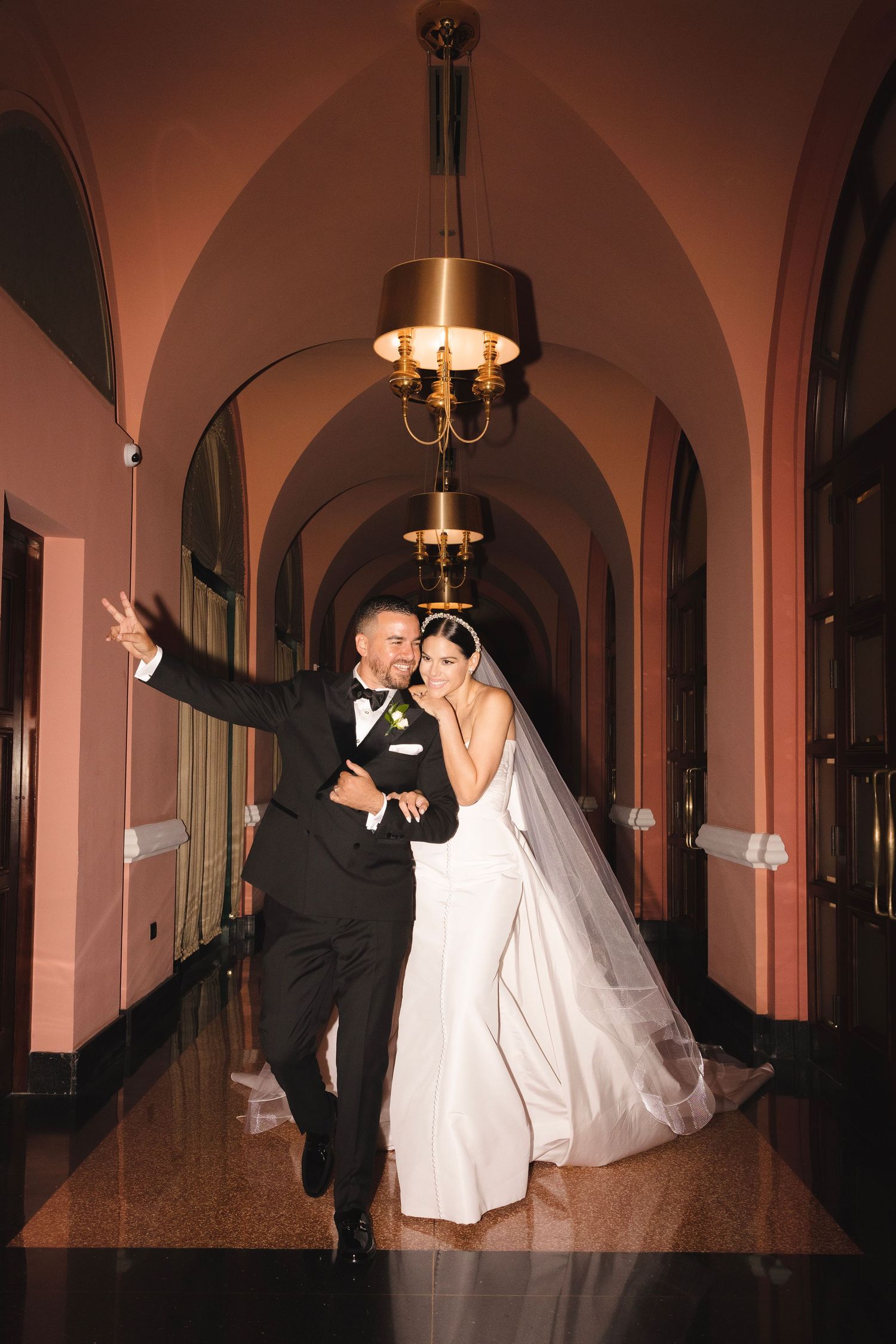 A joyful couple in wedding attire dance and celebrate in an elegant arched hallway with vintage chandeliers overhead.