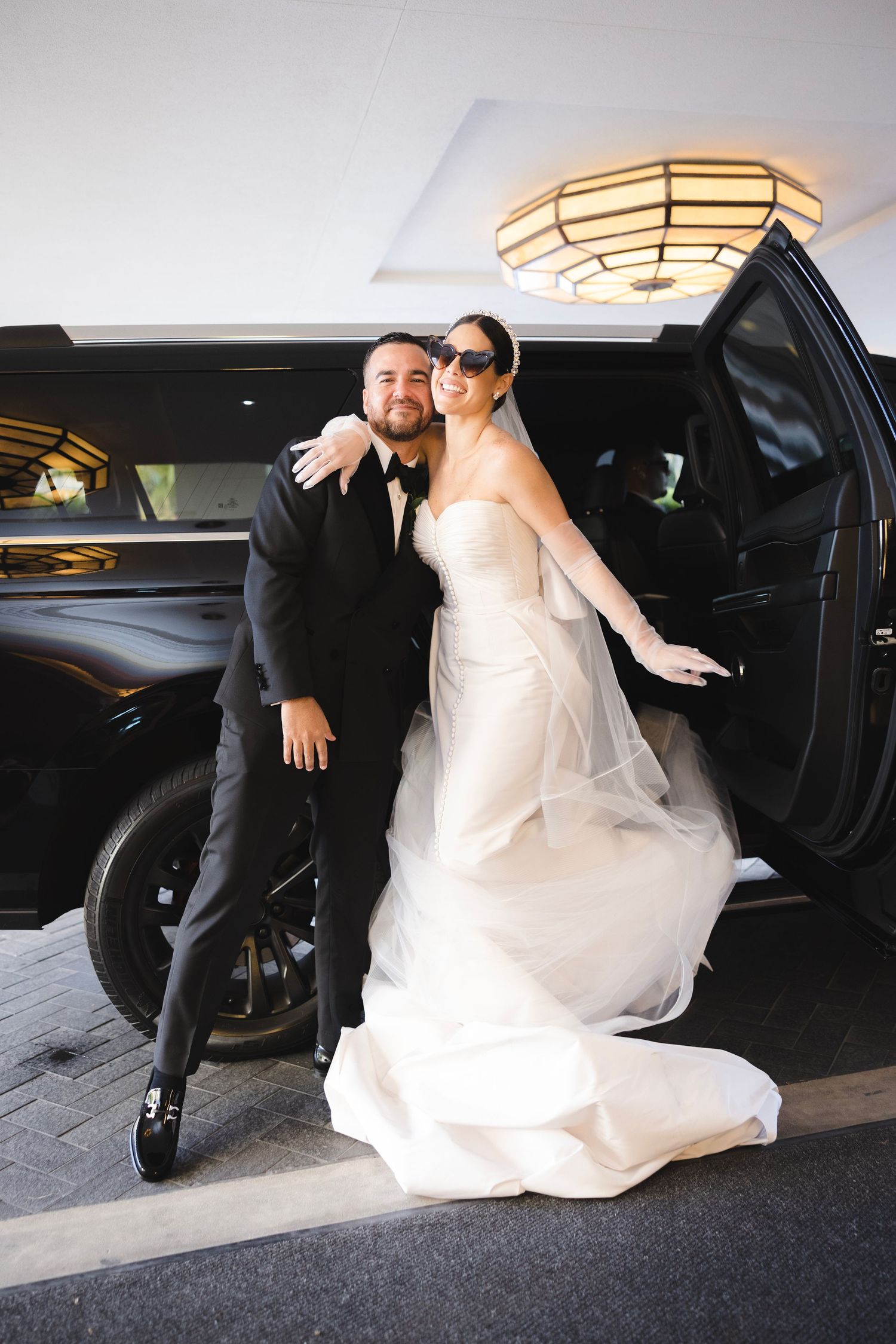 A couple in wedding attire stepping out of a black luxury vehicle under an elegant ceiling light fixture.