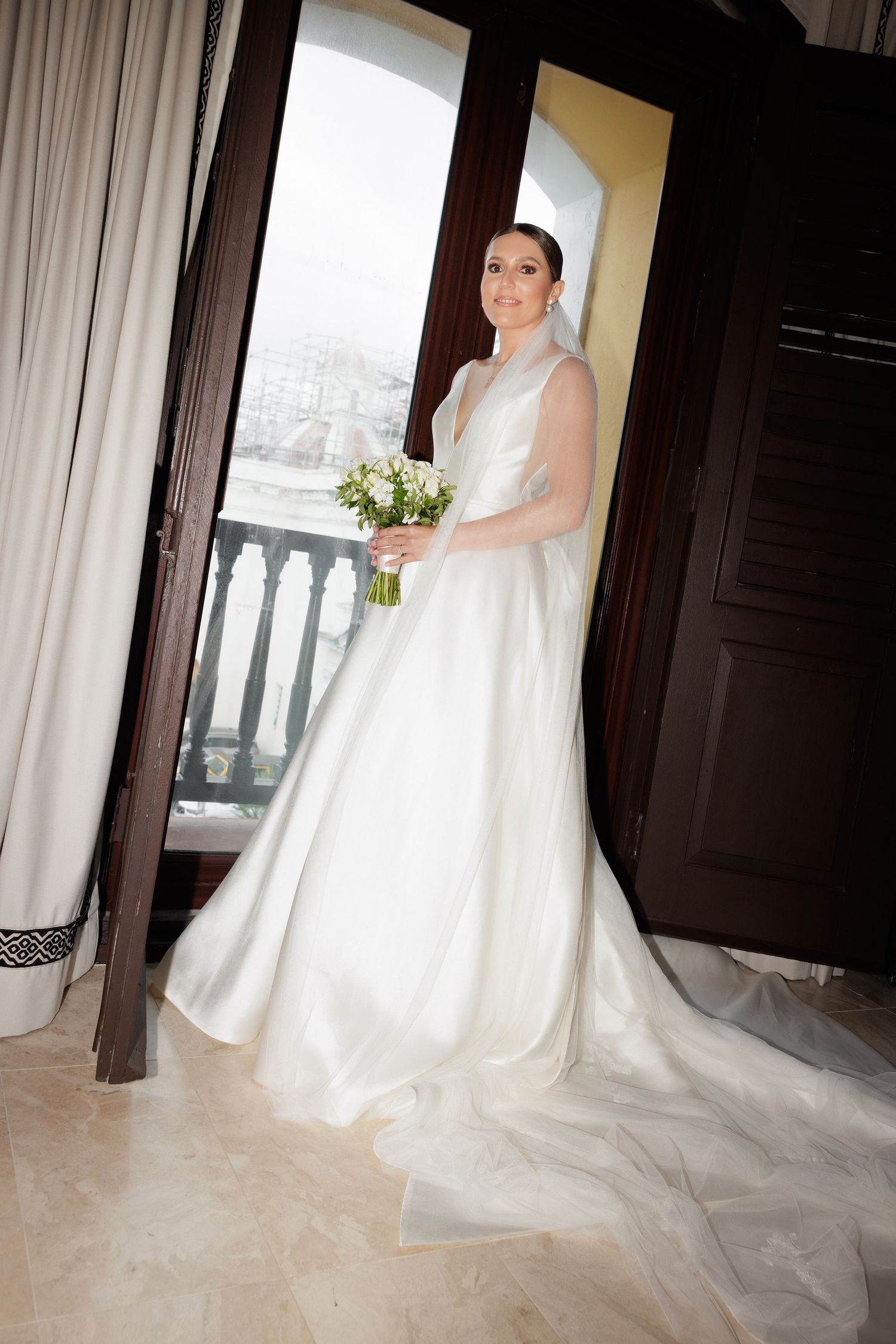 A bride in an elegant white wedding gown with a flowing train stands gracefully against light curtains in an indoor setting.