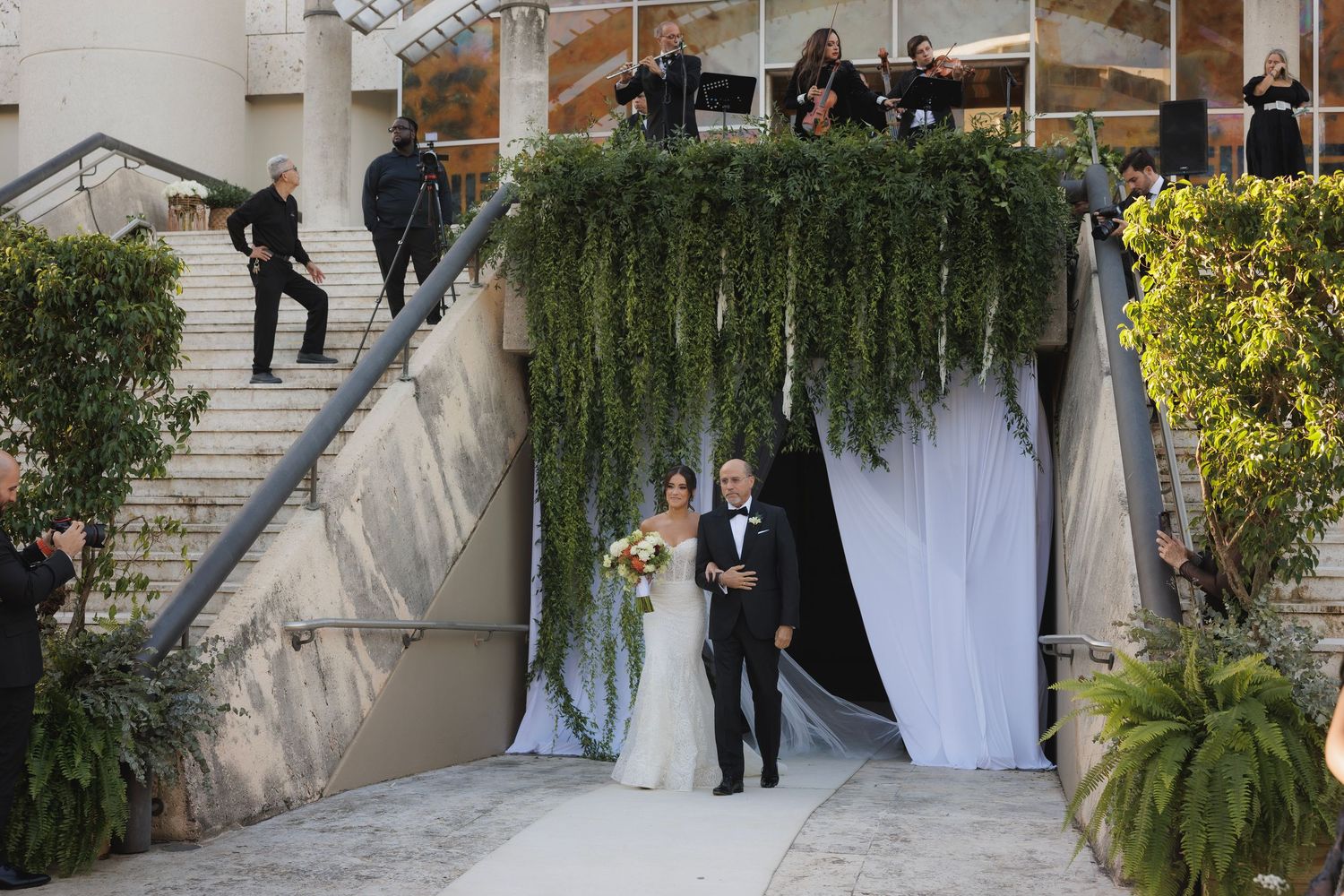 Wedding ceremony guests gathered on elegant stone steps decorated with lush greenery and white drapery.