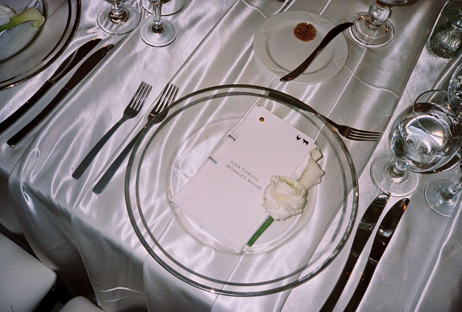 Elegant formal place setting with clear glass plate, white rose, menu card, silverware, and crystal glasses on satin tablecloth.