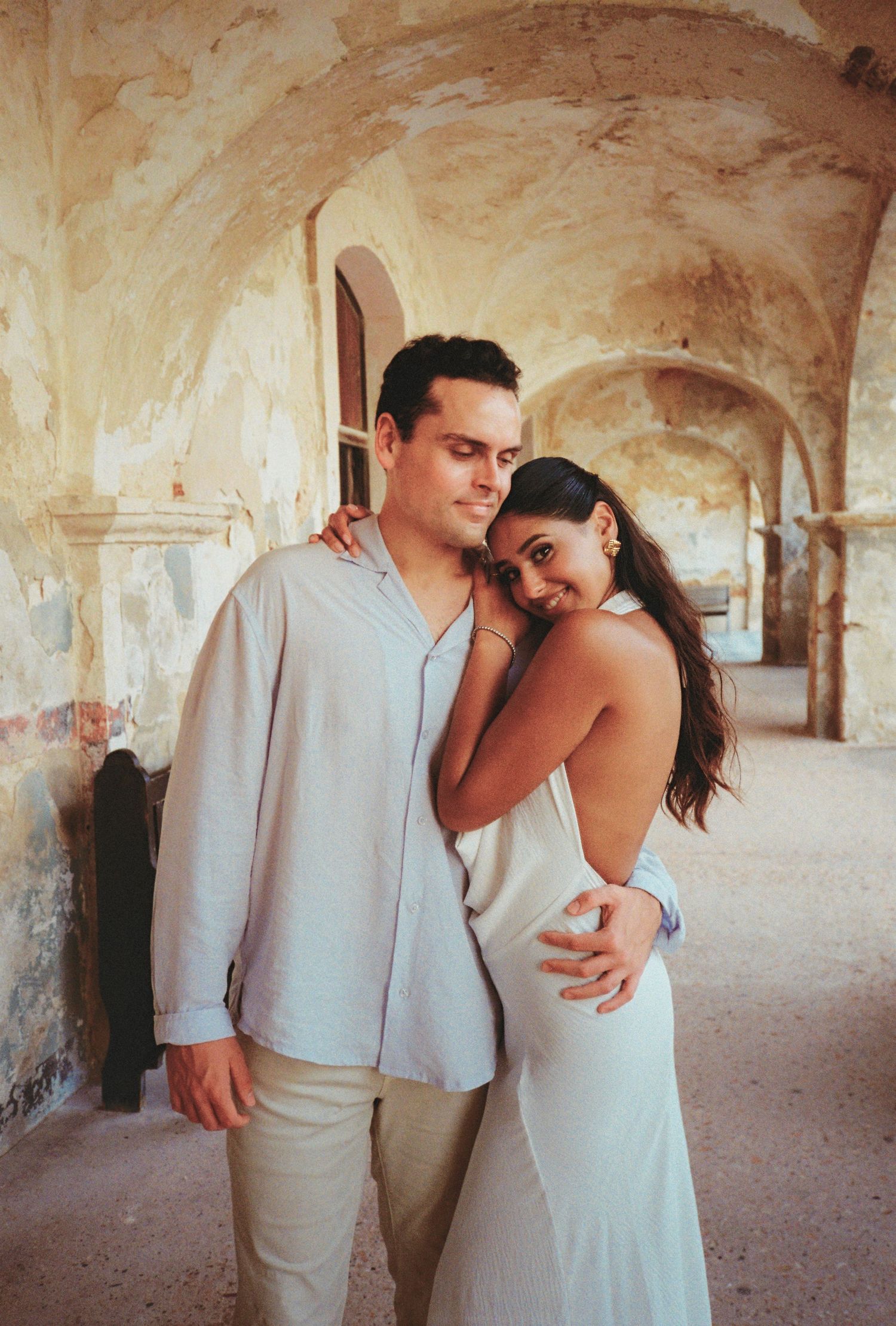 Couple embracing in a rustic arched corridor, woman in backless white dress leaning on mans shoulder.