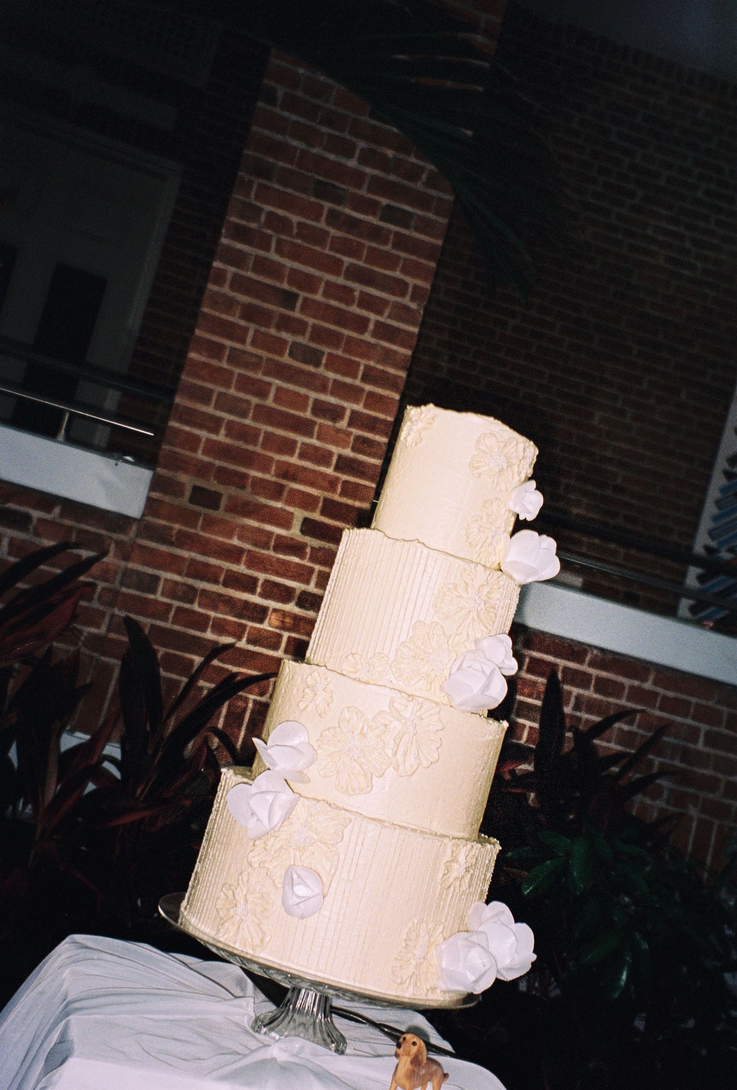 Elegant four-tier white wedding cake adorned with floral decorations displayed outdoors against a brick wall.