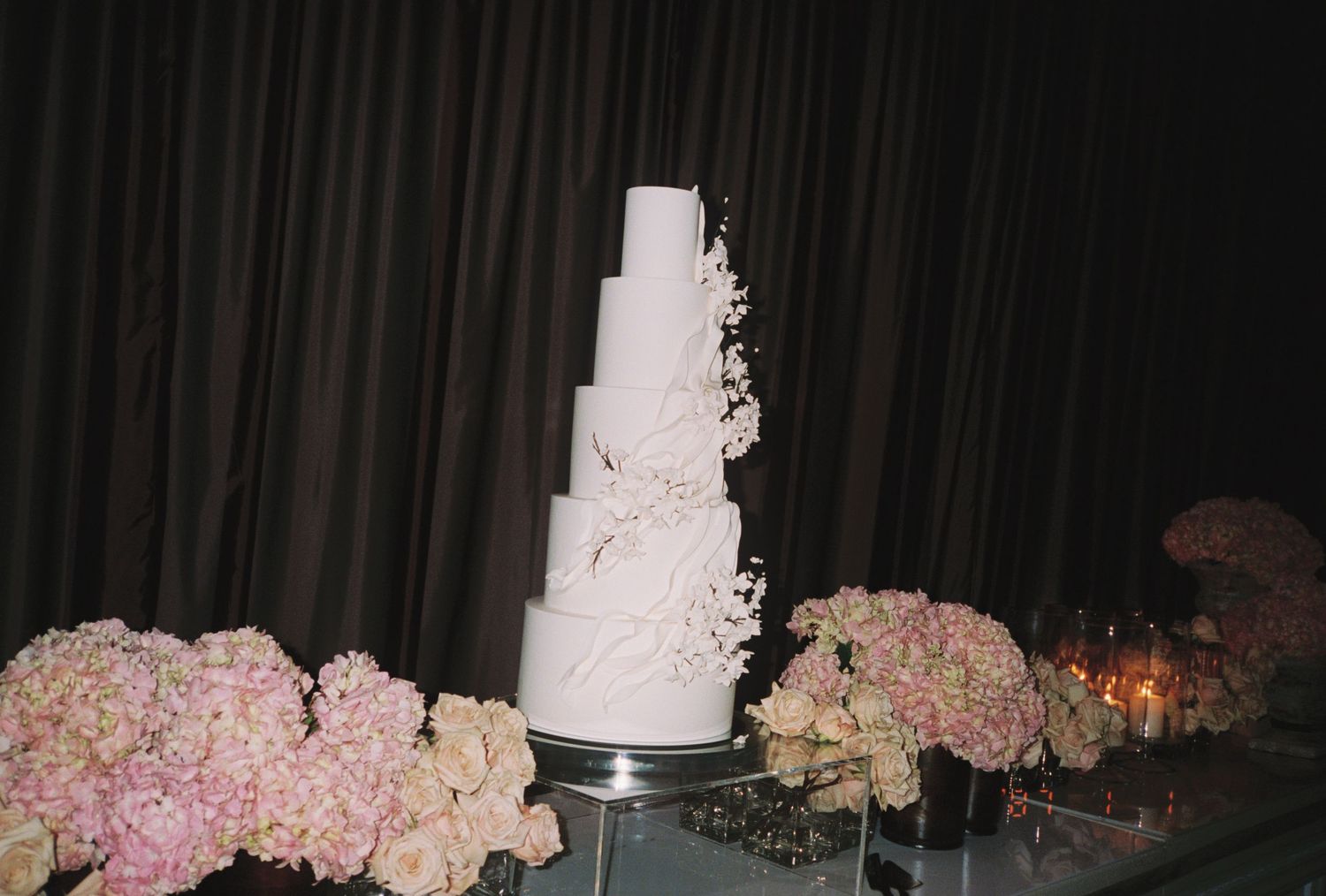 Elegant five-tier white wedding cake adorned with floral details, surrounded by pink hydrangea arrangements on a mirrored table.