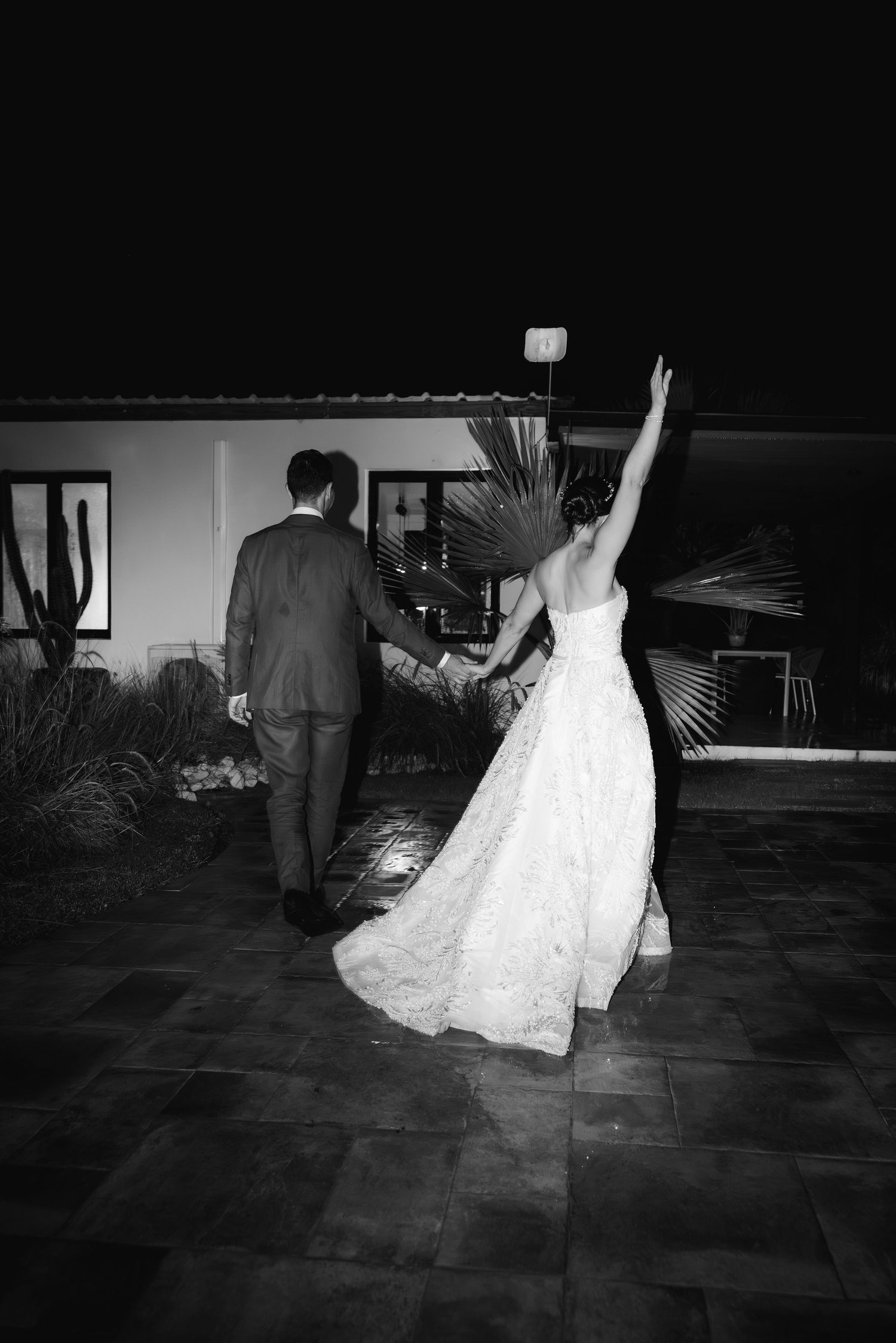 A bride in a white wedding dress waves while walking with her groom on a dark patio at night.