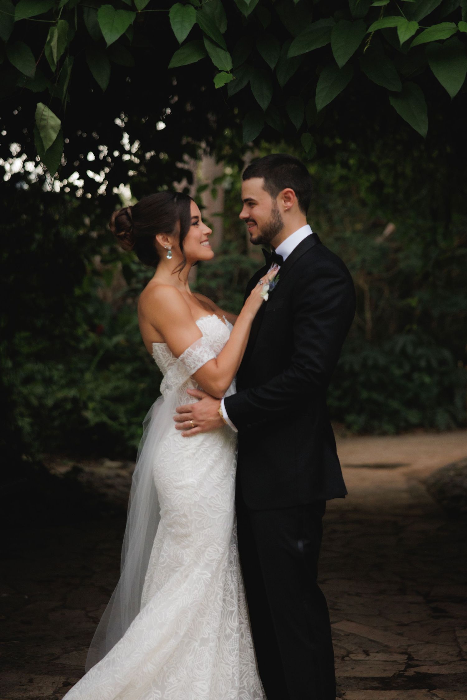 Romantic wedding couple embraces under lush green foliage in an outdoor garden setting.