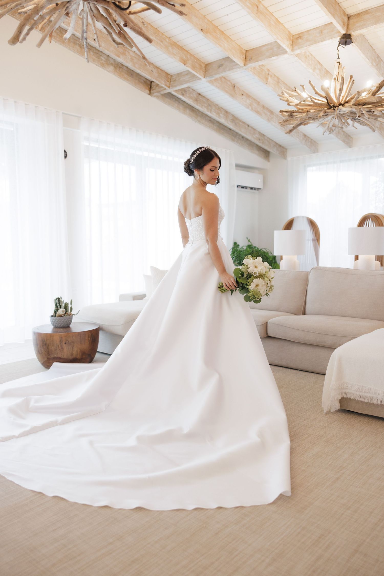 Bride in flowing white gown poses in modern room with rustic wooden beams.