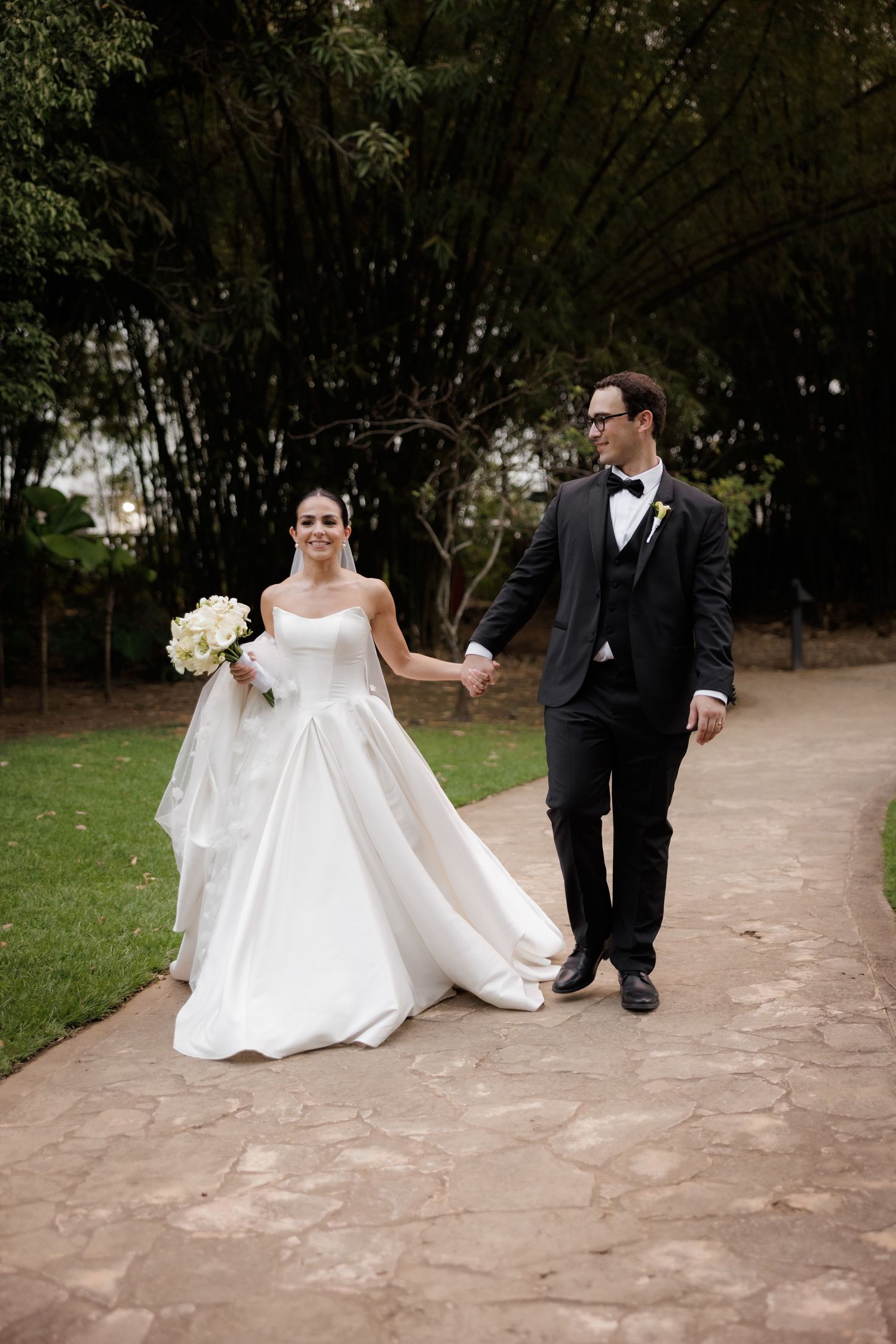 A bride and groom walk together on a stone path during their wedding photoshoot in a garden setting.