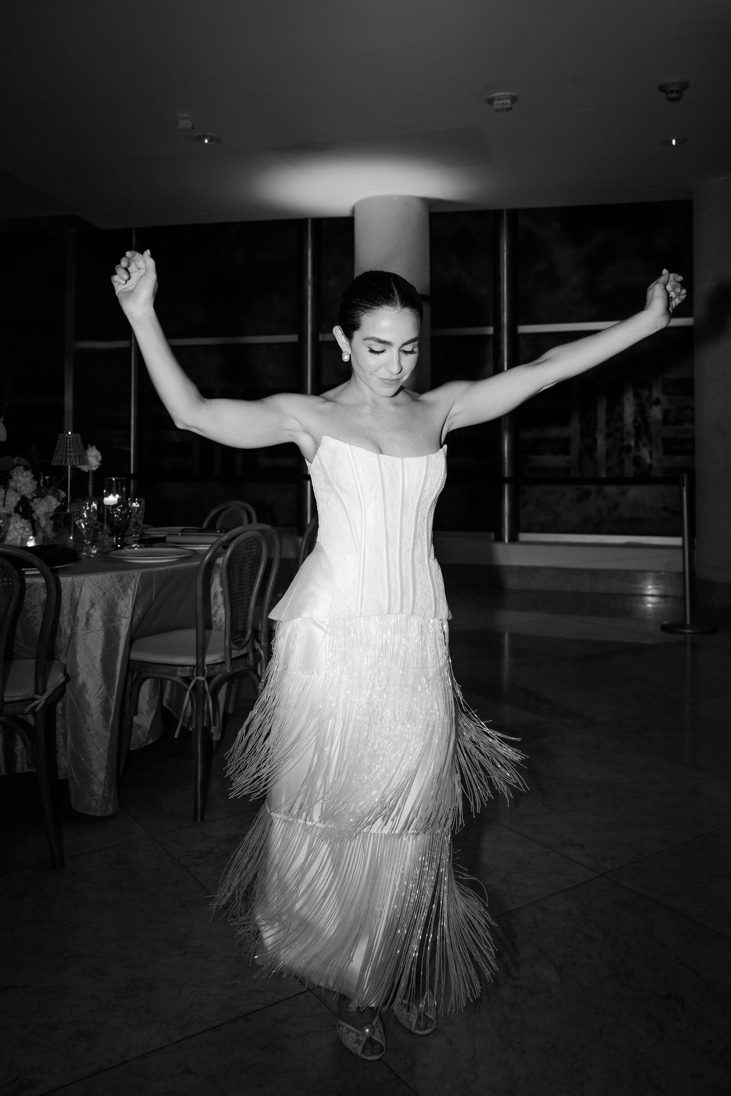 A bride in a white fringe dress dances with her arms raised during a wedding reception.