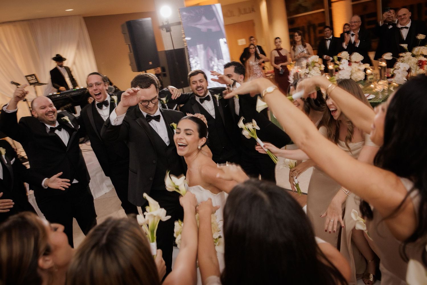 Wedding guests dance and celebrate on a crowded reception dance floor with hands raised in joyful celebration.