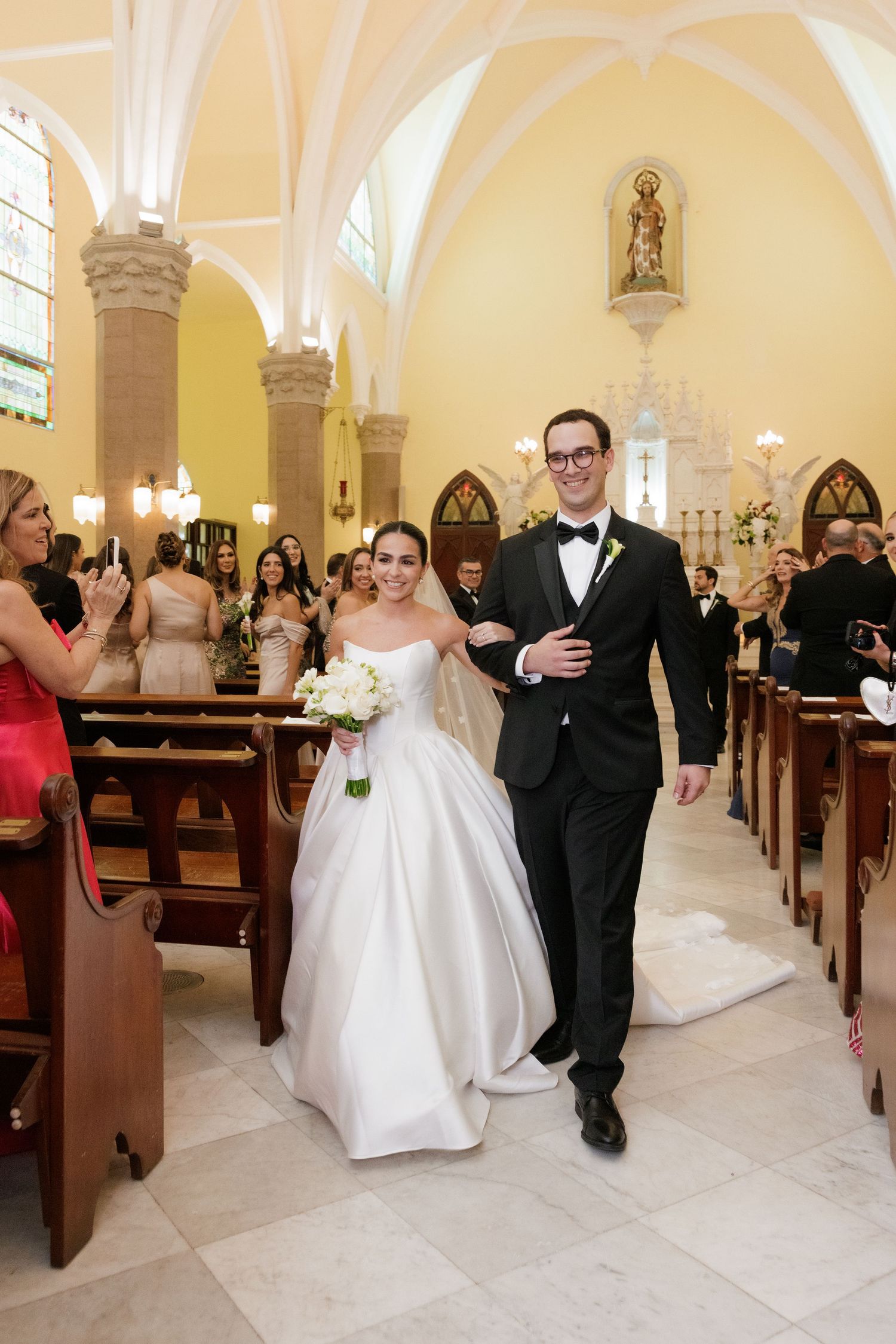 Wedding party members walk down the aisle of a traditional church with wooden pews and vaulted ceilings.