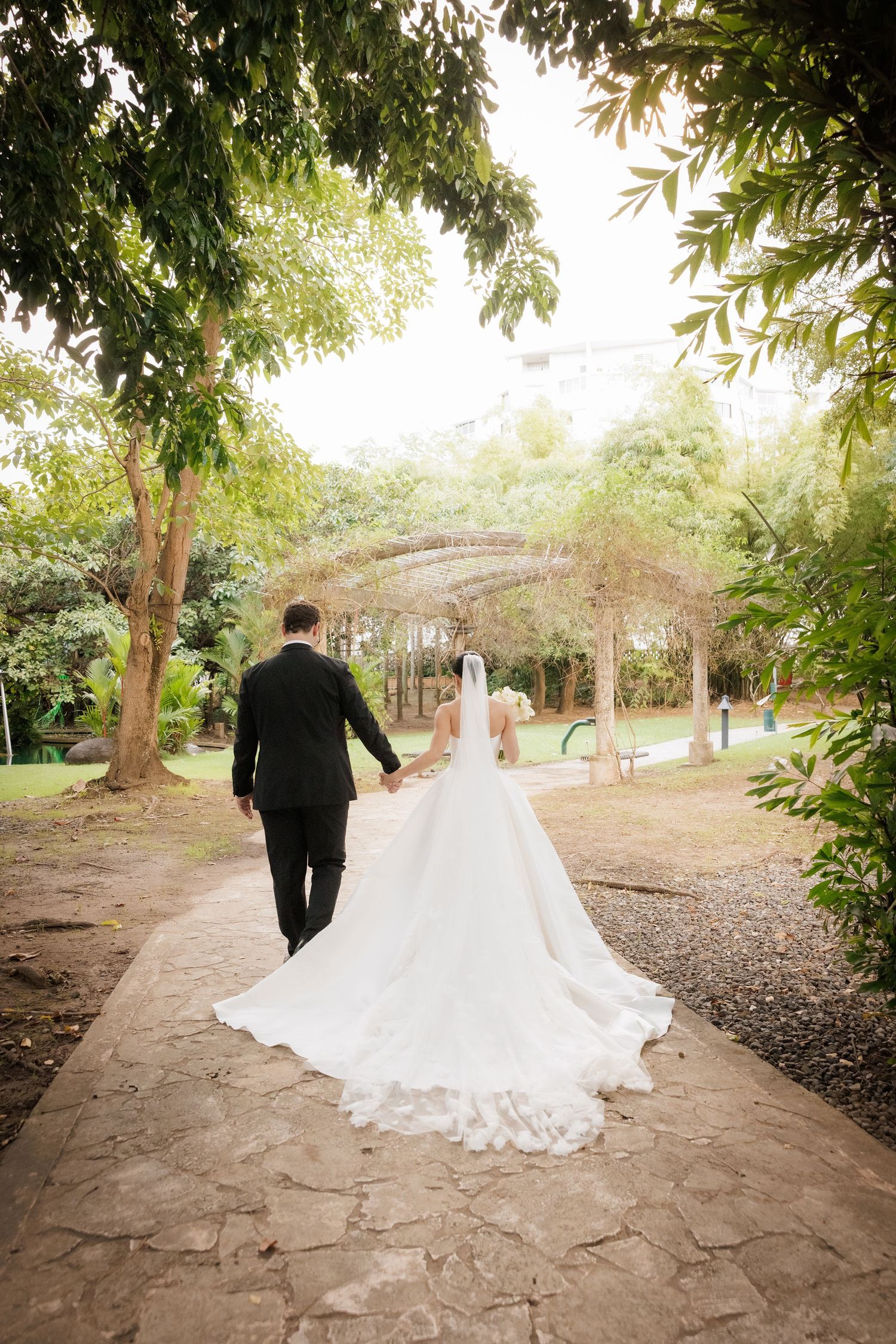 Wedding couple walks along stone path under overhanging tree branches.