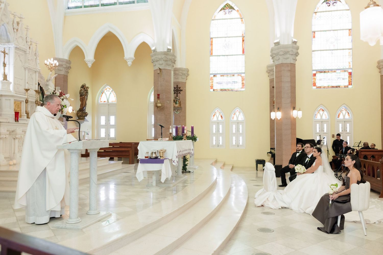 Catholic wedding ceremony in progress with priest at altar and guests seated in ornate church interior.