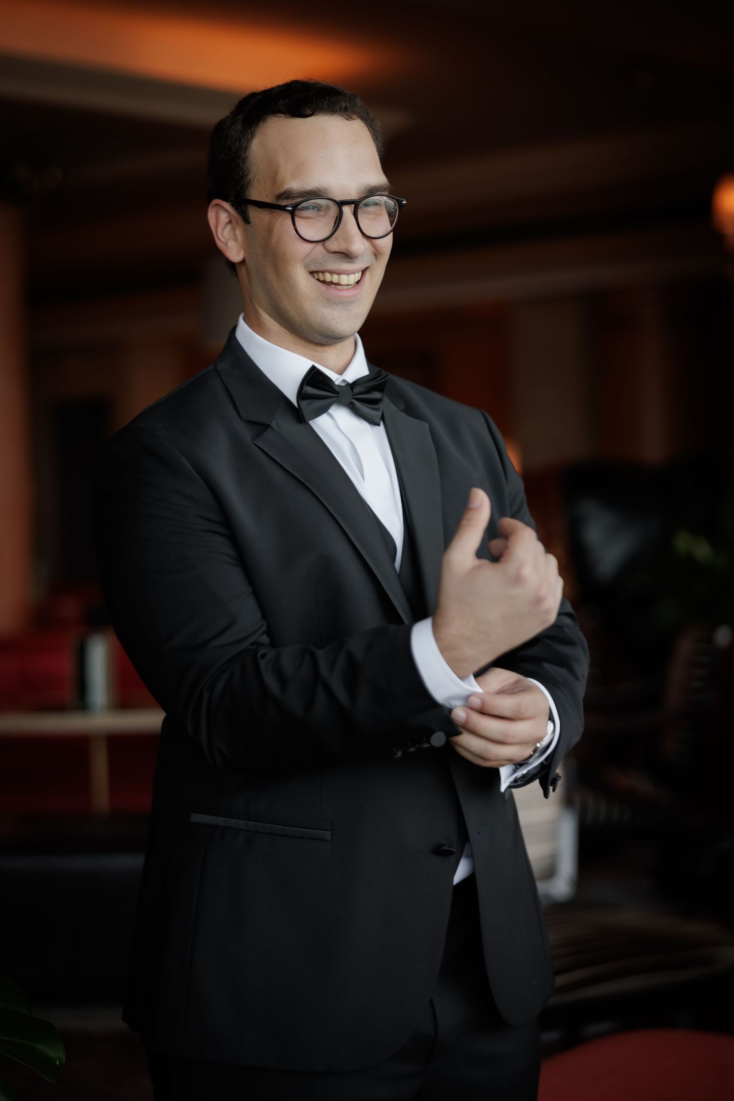 A groom in a black tuxedo and glasses smiles while adjusting his bowtie.