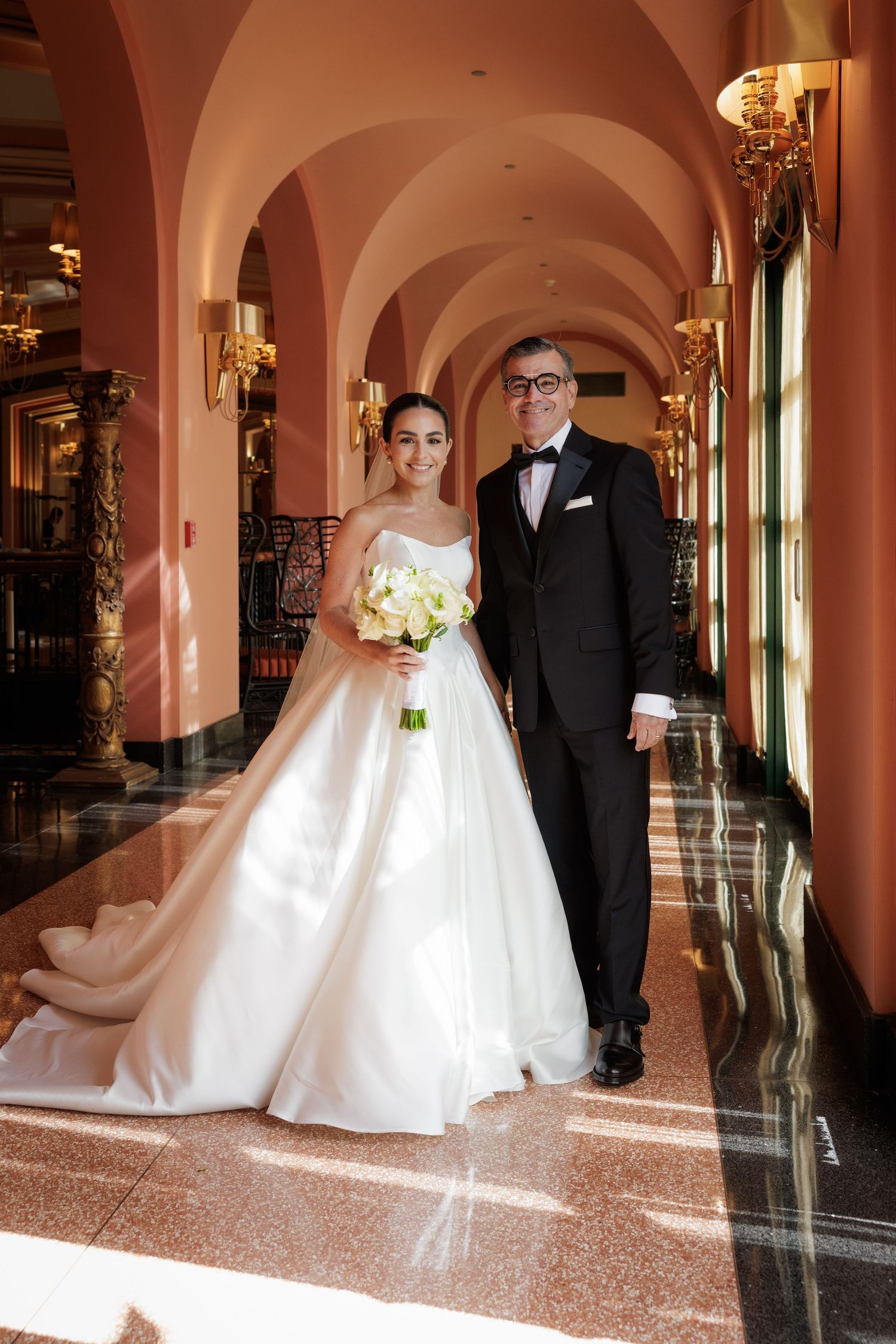Elegant wedding couple poses in a pink-hued hallway with arched columns and marble floors.
