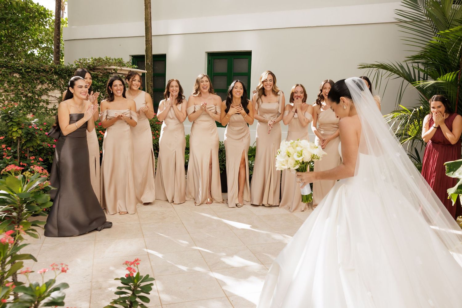 Wedding party in blush pink bridesmaid dresses poses outdoors at elegant garden venue.