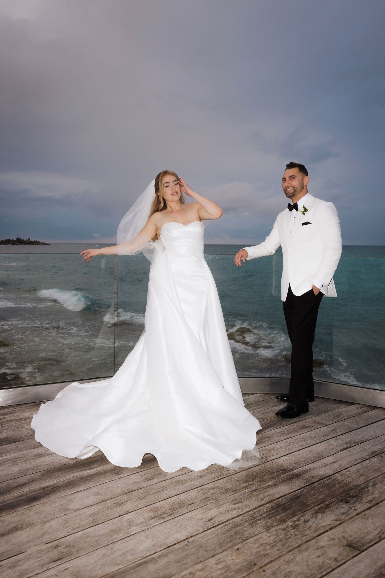 A couple poses for wedding photos on a wooden deck overlooking turquoise waters and stormy skies.