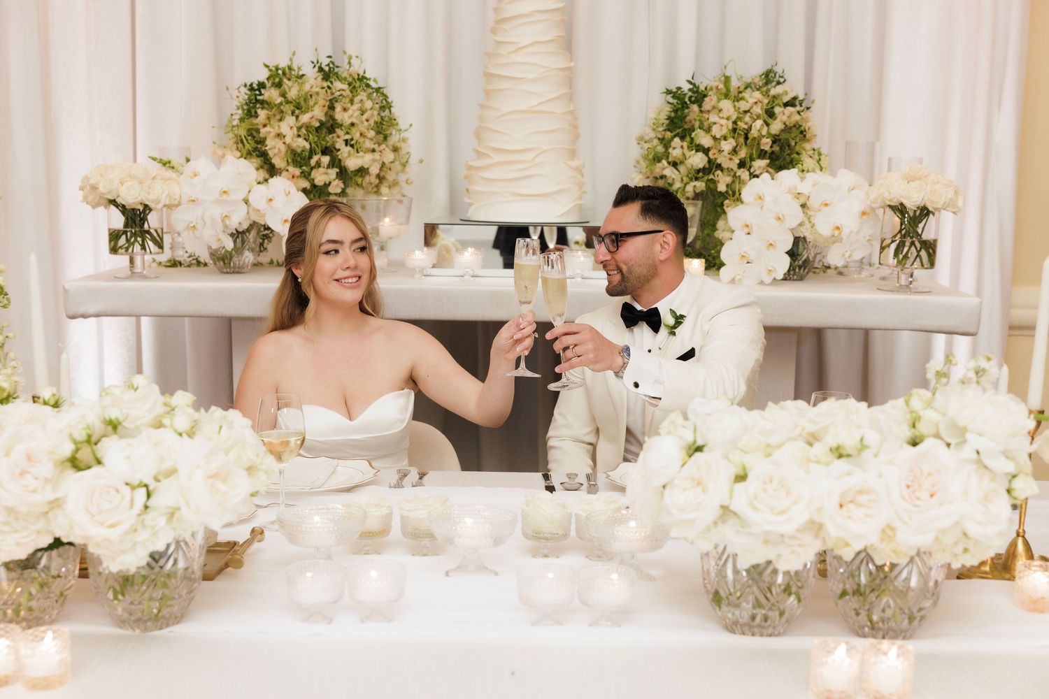Wedding reception tables decorated with elegant white floral centerpieces and crystal chandeliers in a luxurious ballroom.