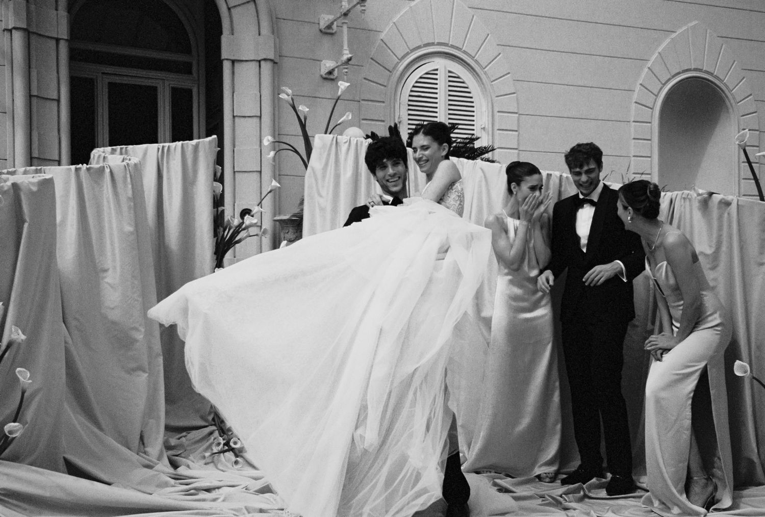 Black and white photo of a bride in a flowing white gown surrounded by attendants outside an elegant building.