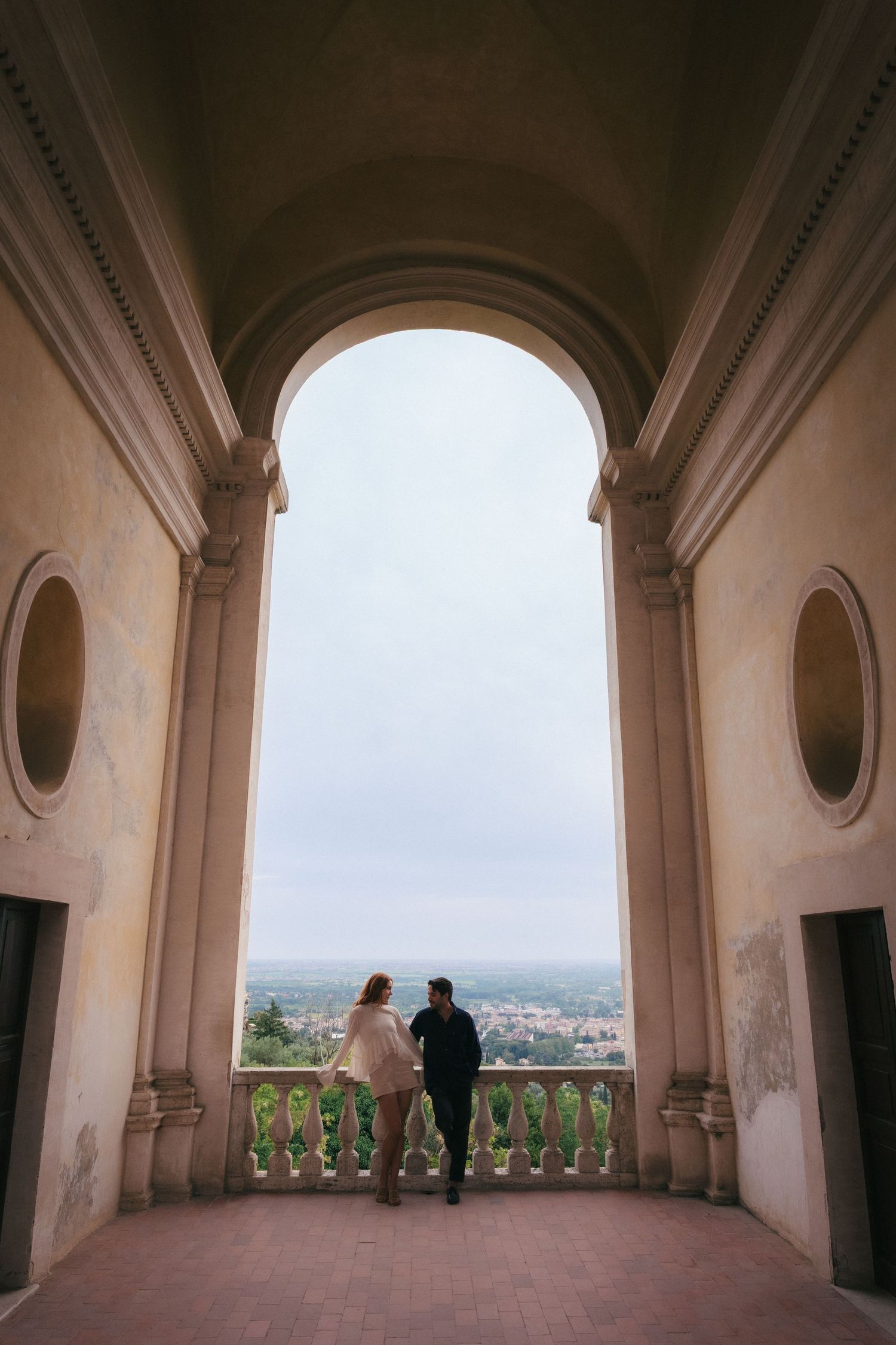 Couple standing on elegant arched balcony overlooking city panorama at historic Italian villa.