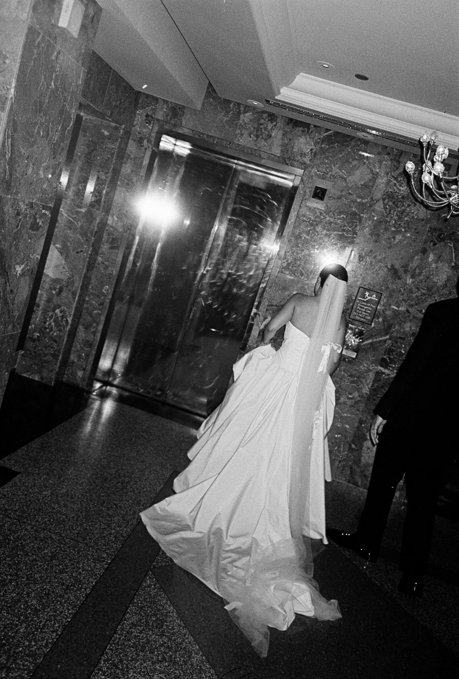 Black and white photo of a bride in a flowing white gown walking toward elevator doors in an elegant marble lobby.