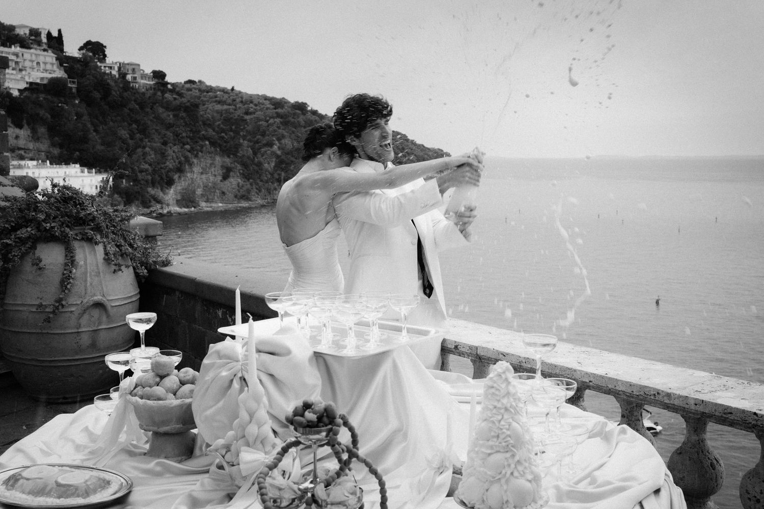 Black and white photo of a bride and groom celebrating on a seaside terrace with champagne glasses and ocean views.