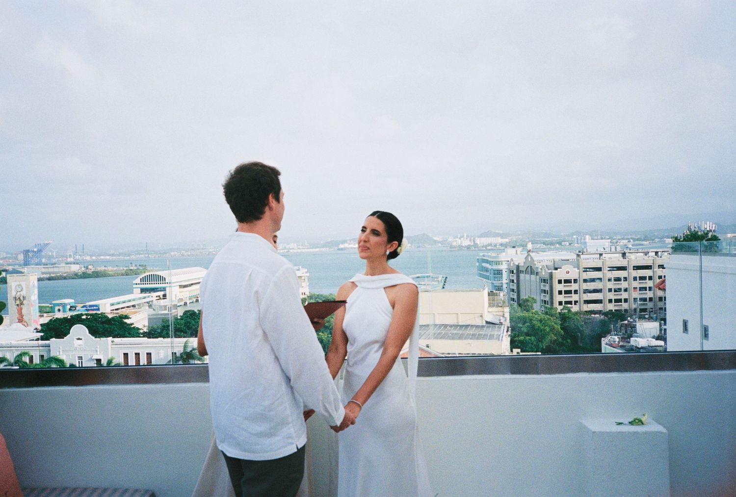 Couple holding hands on rooftop terrace with ocean and city skyline backdrop during outdoor wedding ceremony.