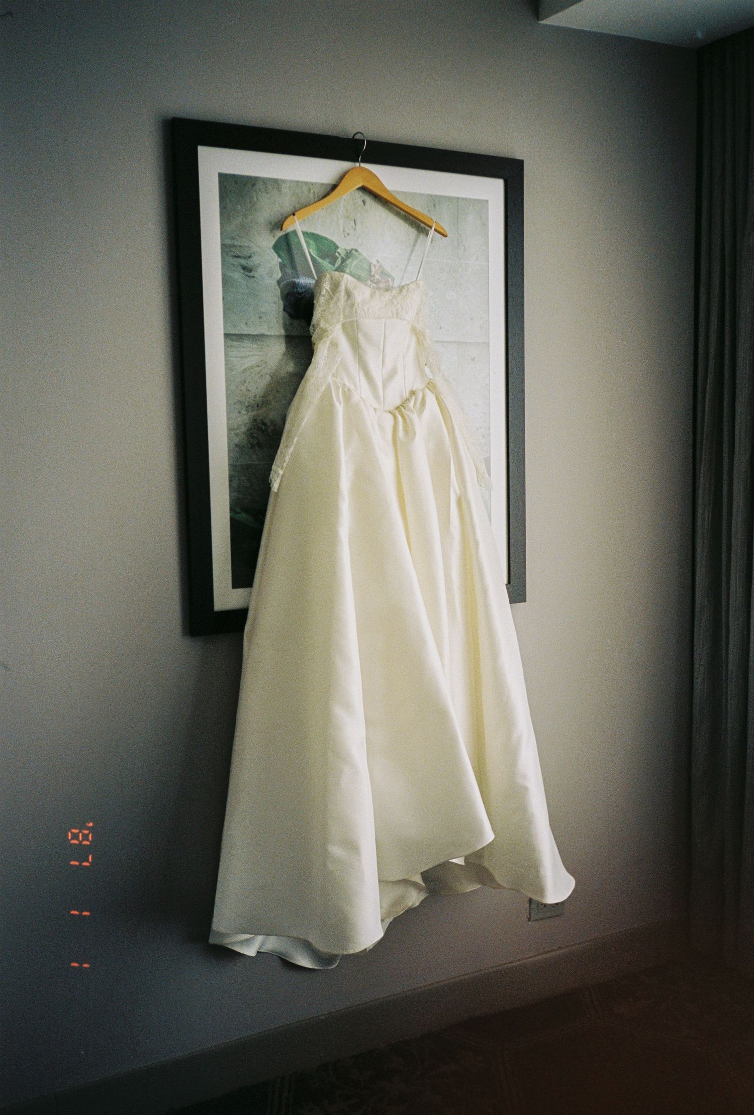 Ivory ball gown wedding dress hanging on wooden hanger in front of framed artwork in hotel room.