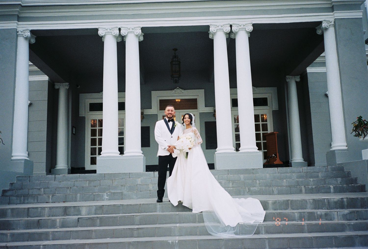 Bride and groom posing on steps of elegant white columned mansion venue on their wedding day.