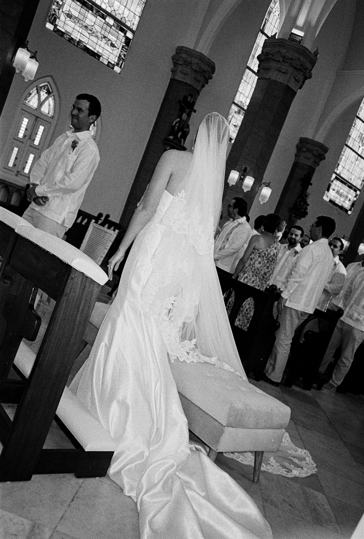 Bride in white gown and veil kneels at altar during church wedding ceremony in black and white photo.