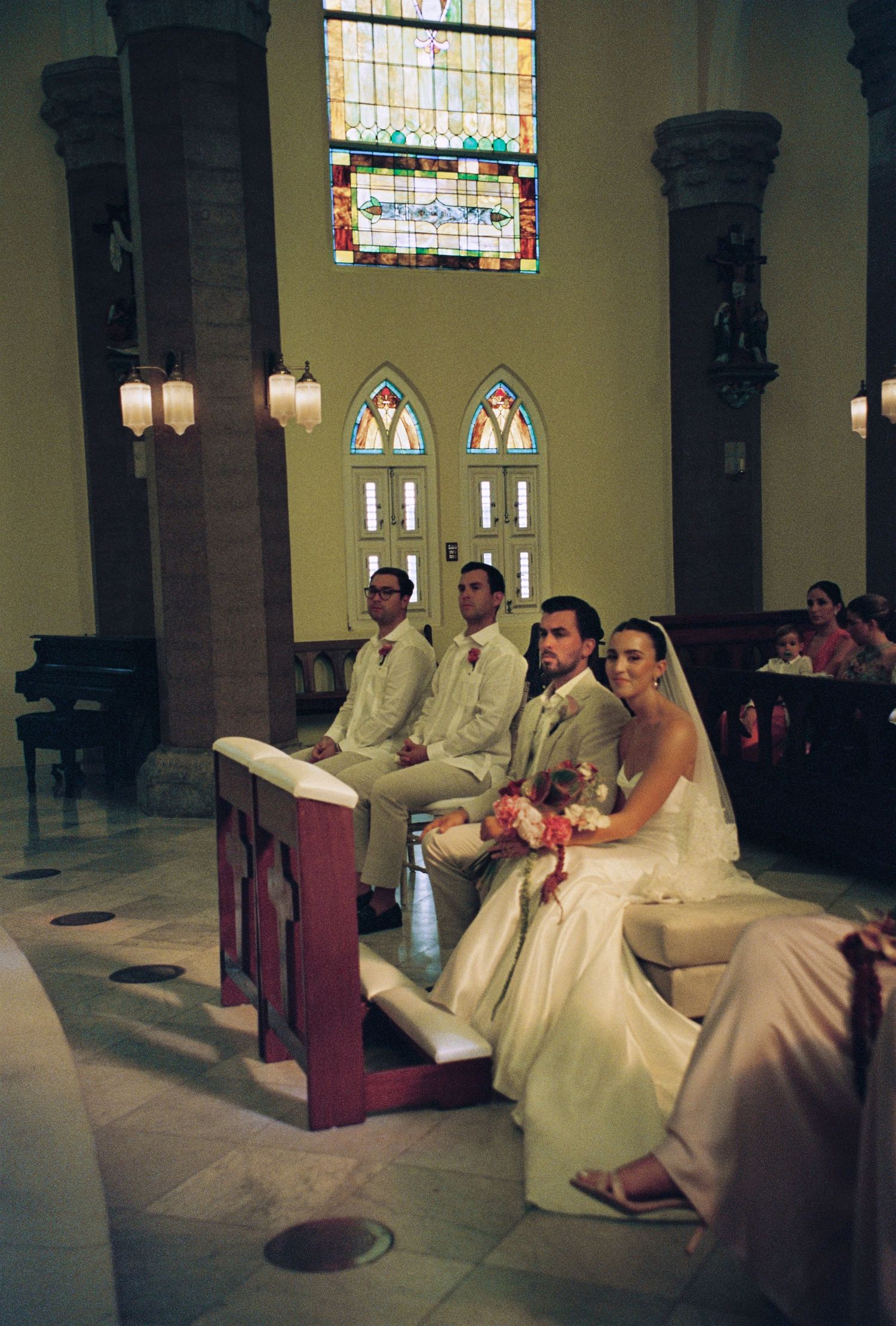 Bride and groom sit at church altar with groomsmen during wedding ceremony with stained glass windows.
