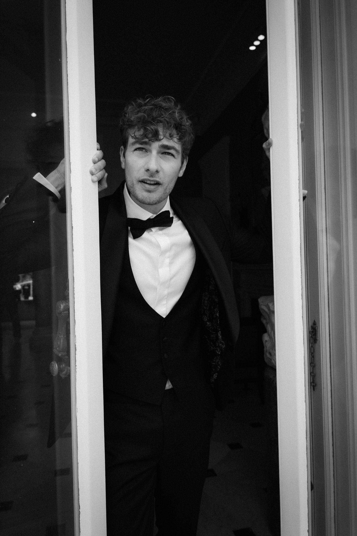 Black and white photo of a young man in a tuxedo with bow tie, posing in a doorway at a formal event.