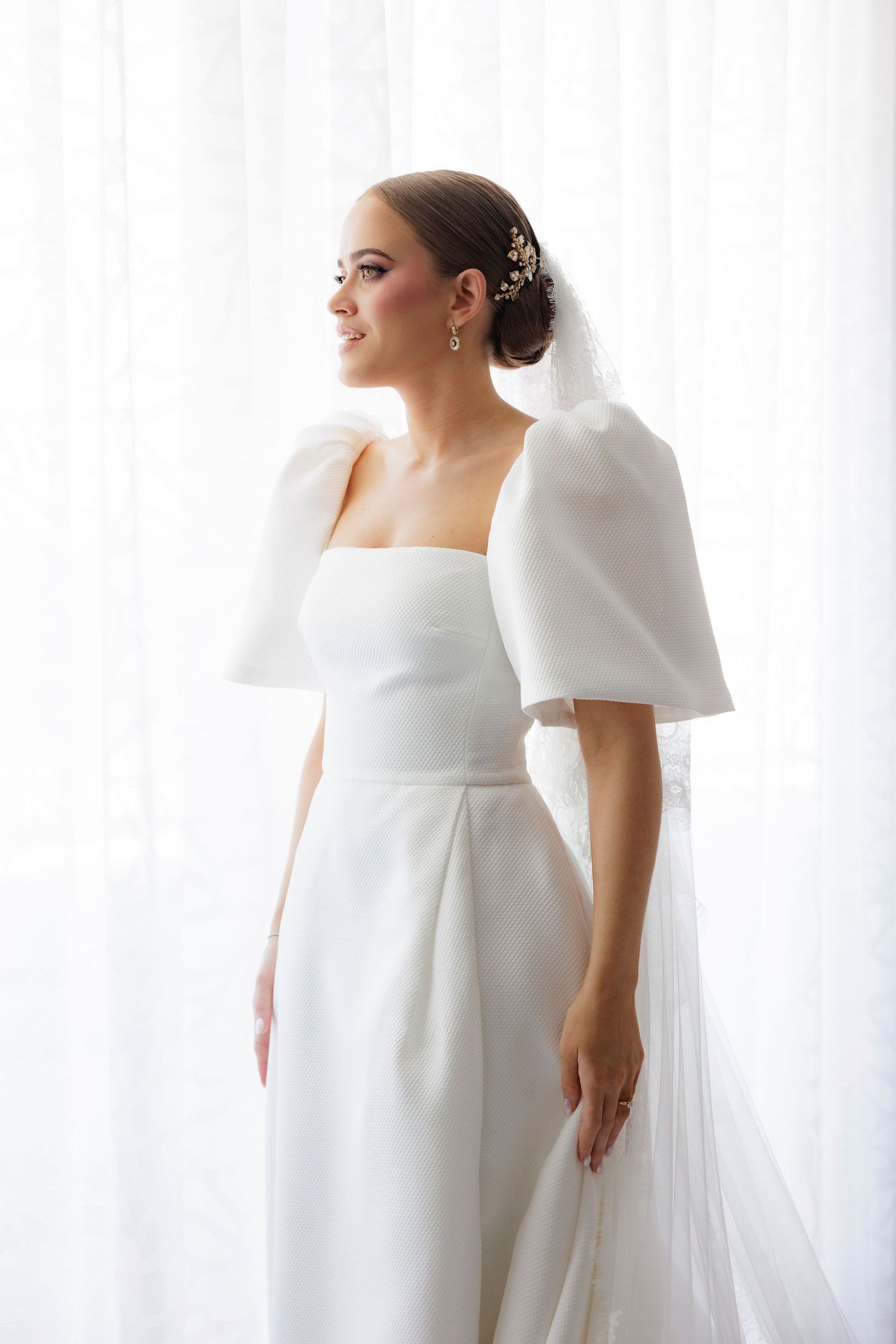 A bride in an elegant white wedding dress with dramatic puff sleeves stands near a window in natural light.