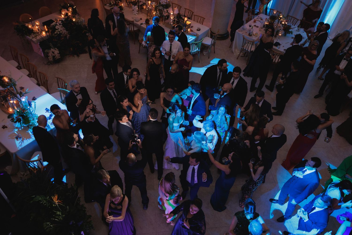 Wedding reception dance floor illuminated with blue lighting as guests gather and celebrate in a dark banquet hall.