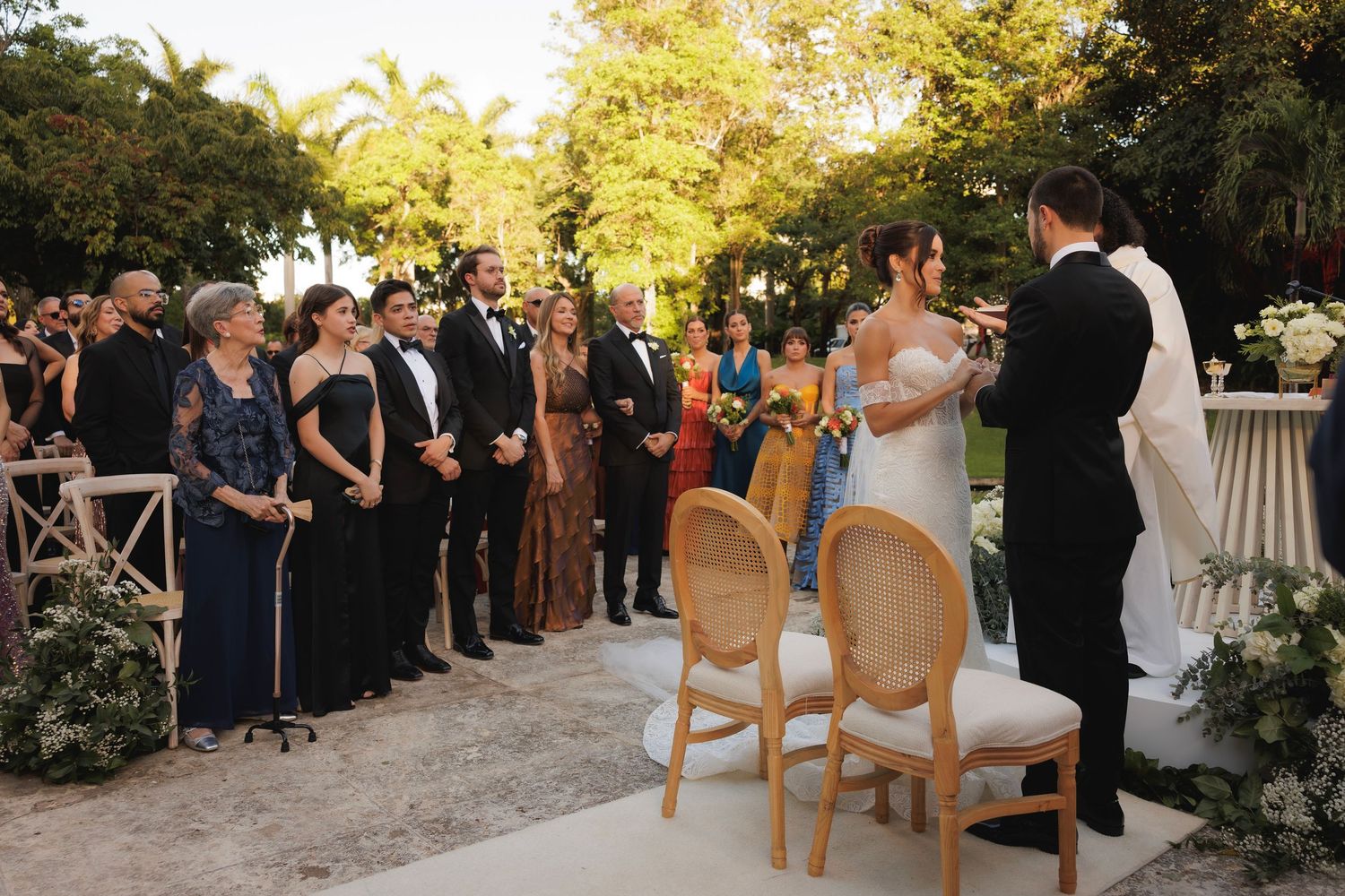 Guests gather outdoors at a sunset wedding ceremony with wooden chairs and white floral decor.