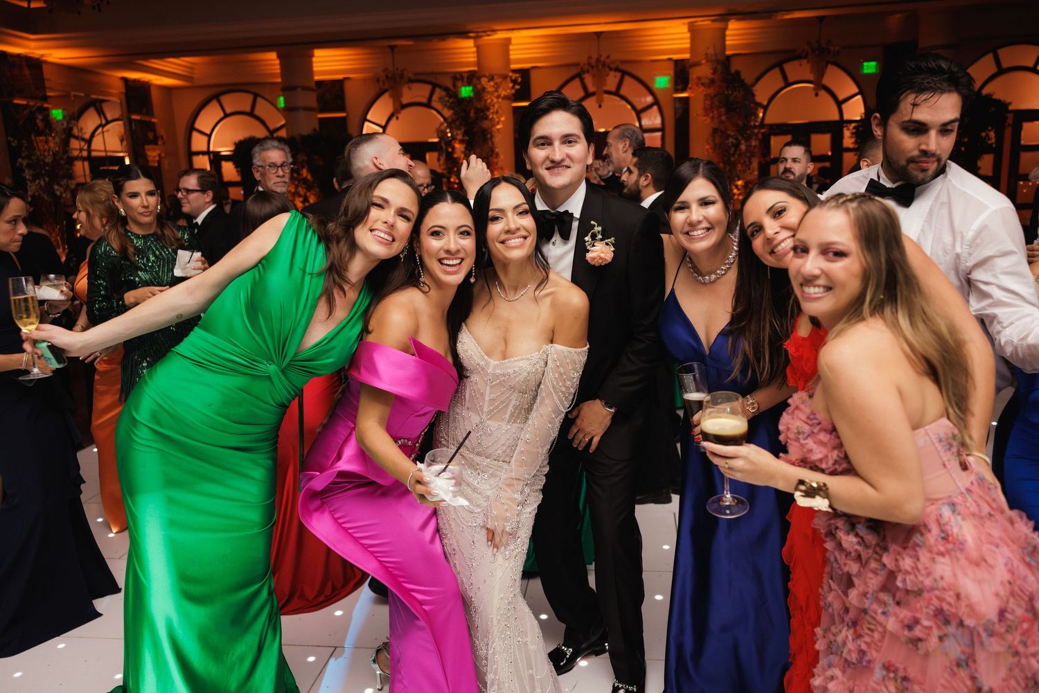 Wedding guests in vibrant formal dresses celebrate and dance together at an elegant reception venue.