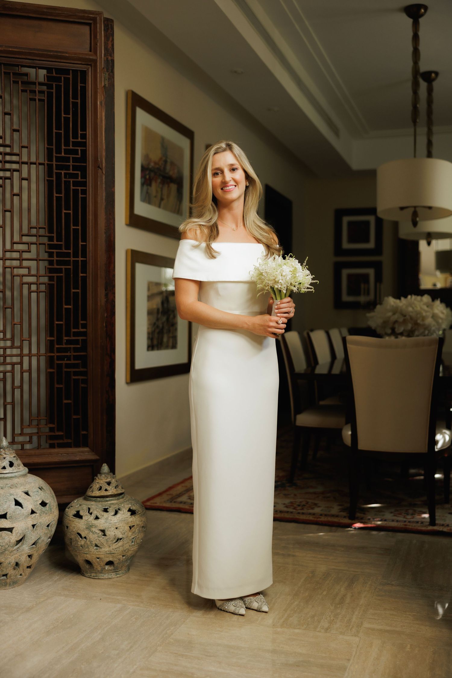 A bride in an elegant off-shoulder white column dress holds a delicate dried flower bouquet in an elegant interior setting.