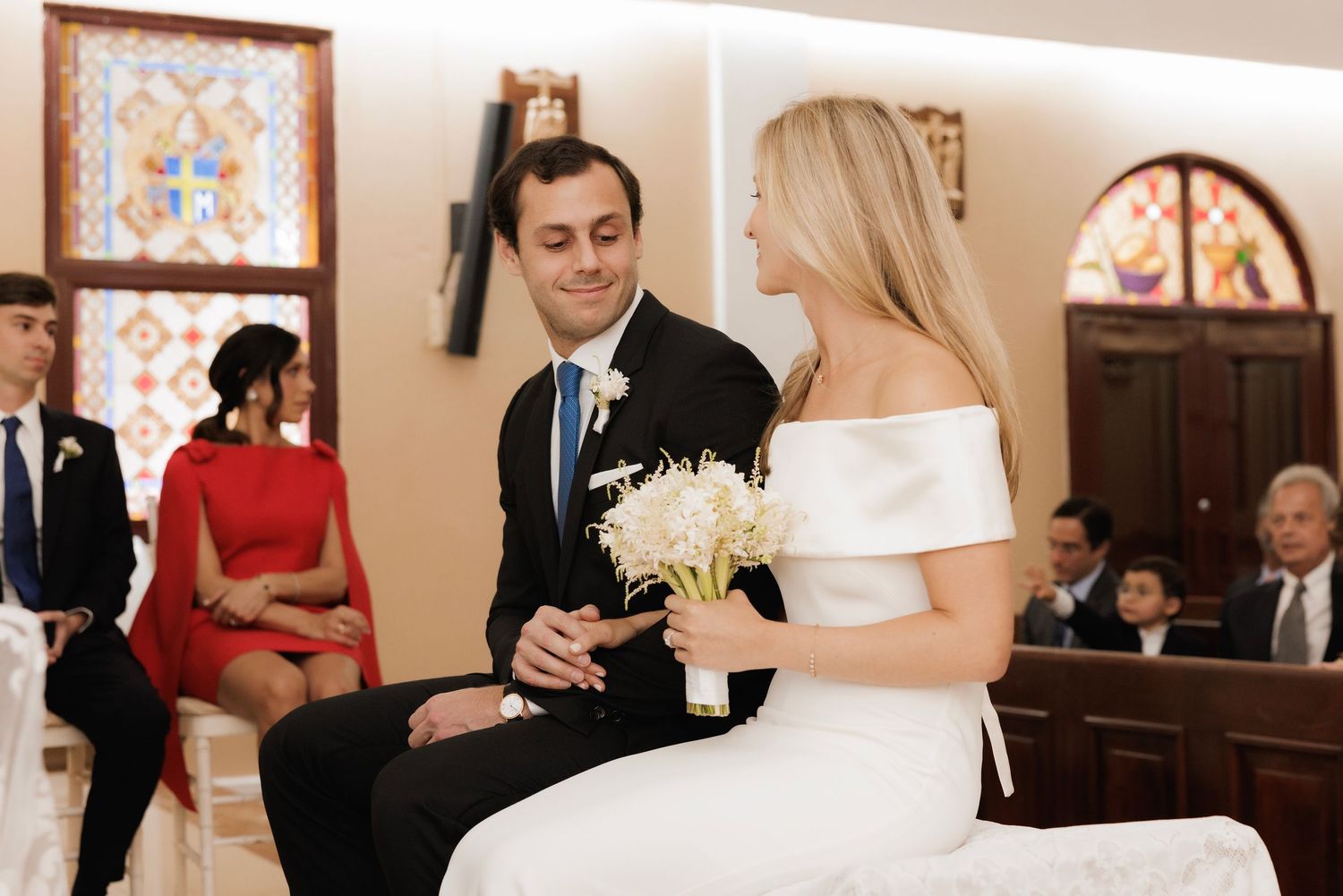 A bride in an elegant off-shoulder white dress sits with her groom during their intimate church wedding ceremony.