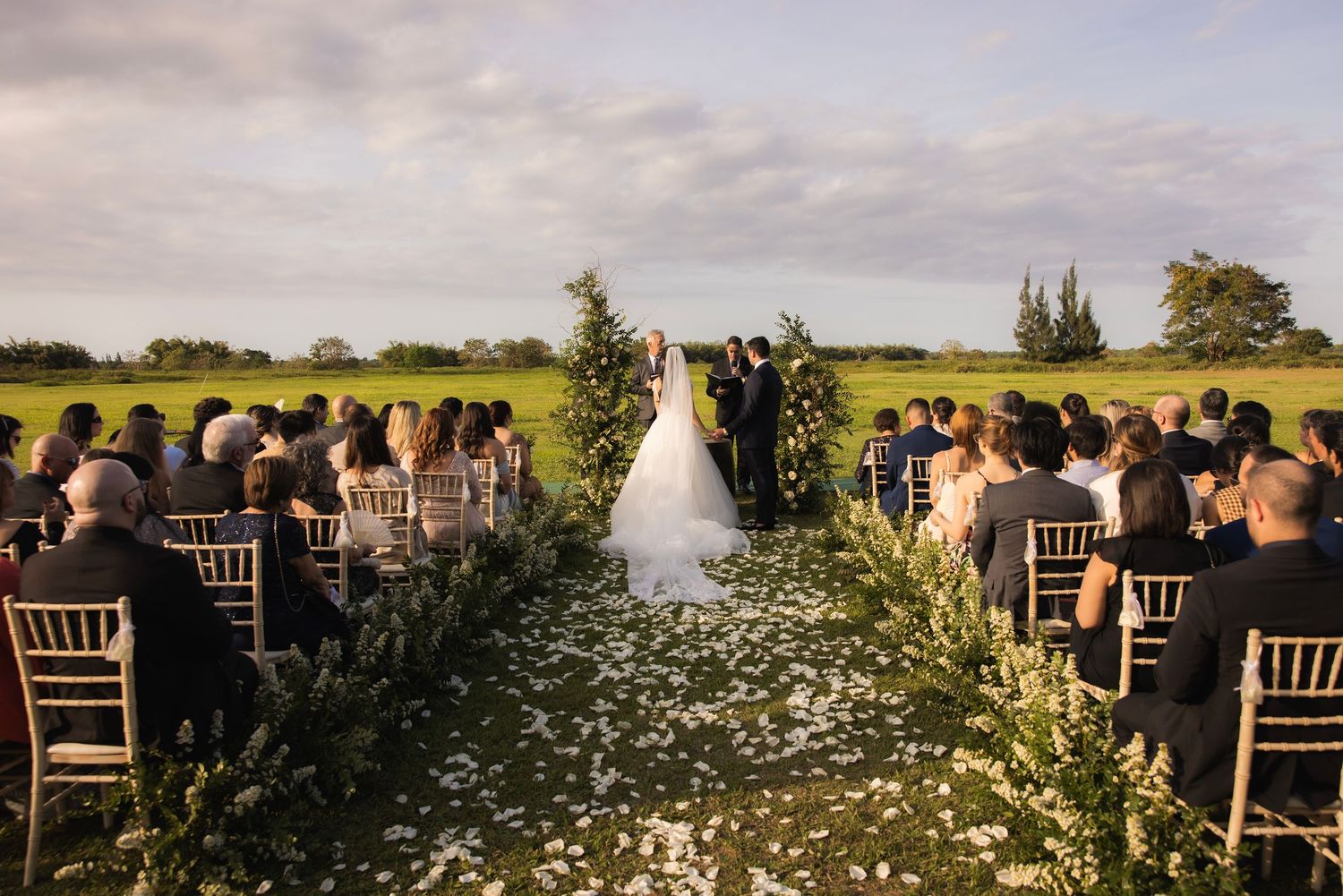 Rustic outdoor wedding ceremony setup with wooden chairs and floral aisle decorations.