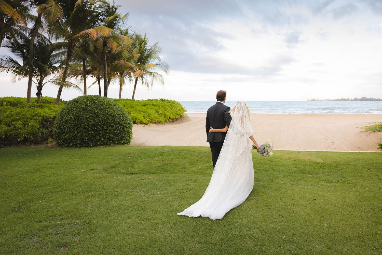 Couple in wedding attire stands on lush green lawn overlooking tropical beach with palm trees and ocean views.