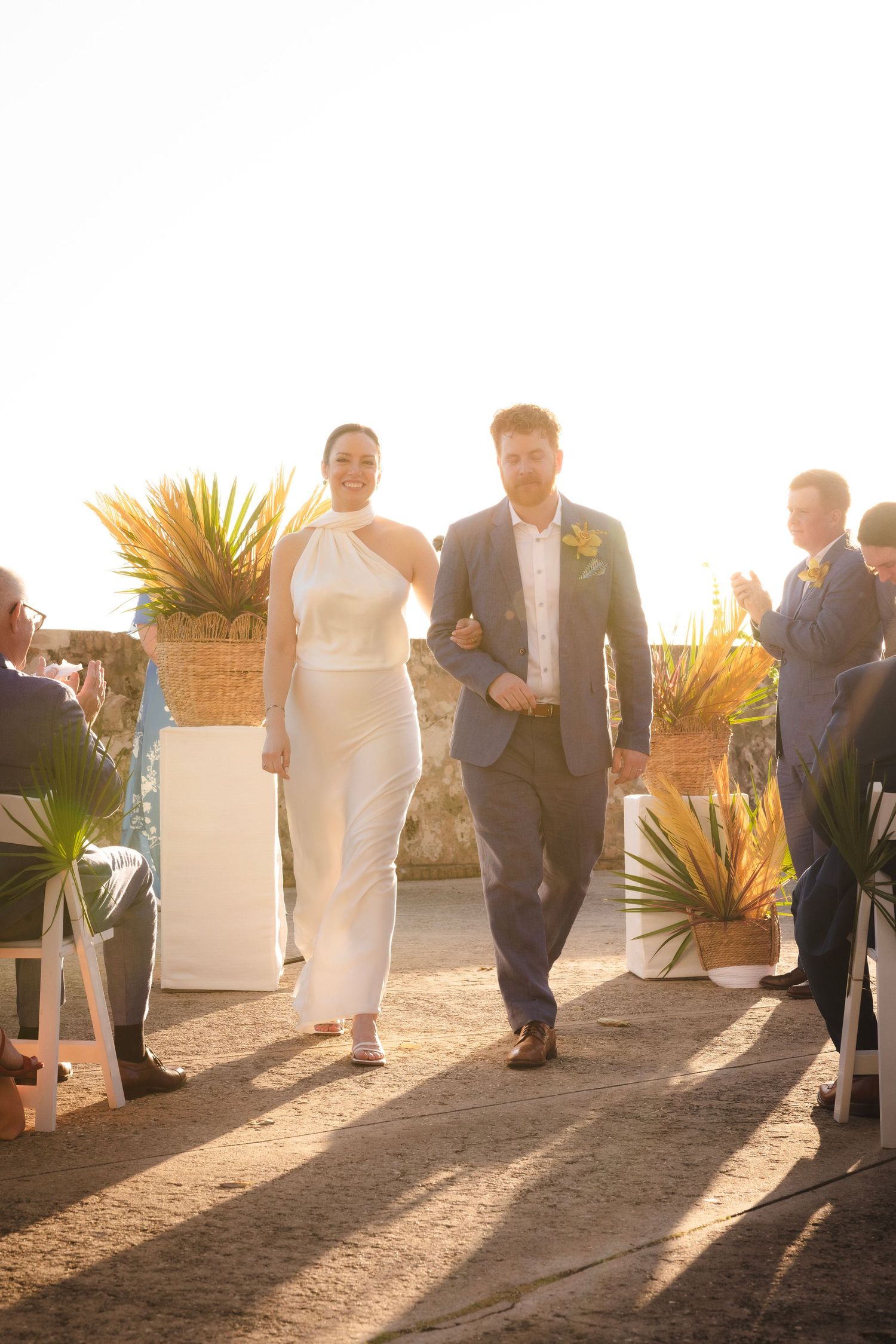 Wedding party poses on a beachfront terrace during golden hour with palm trees in the background.