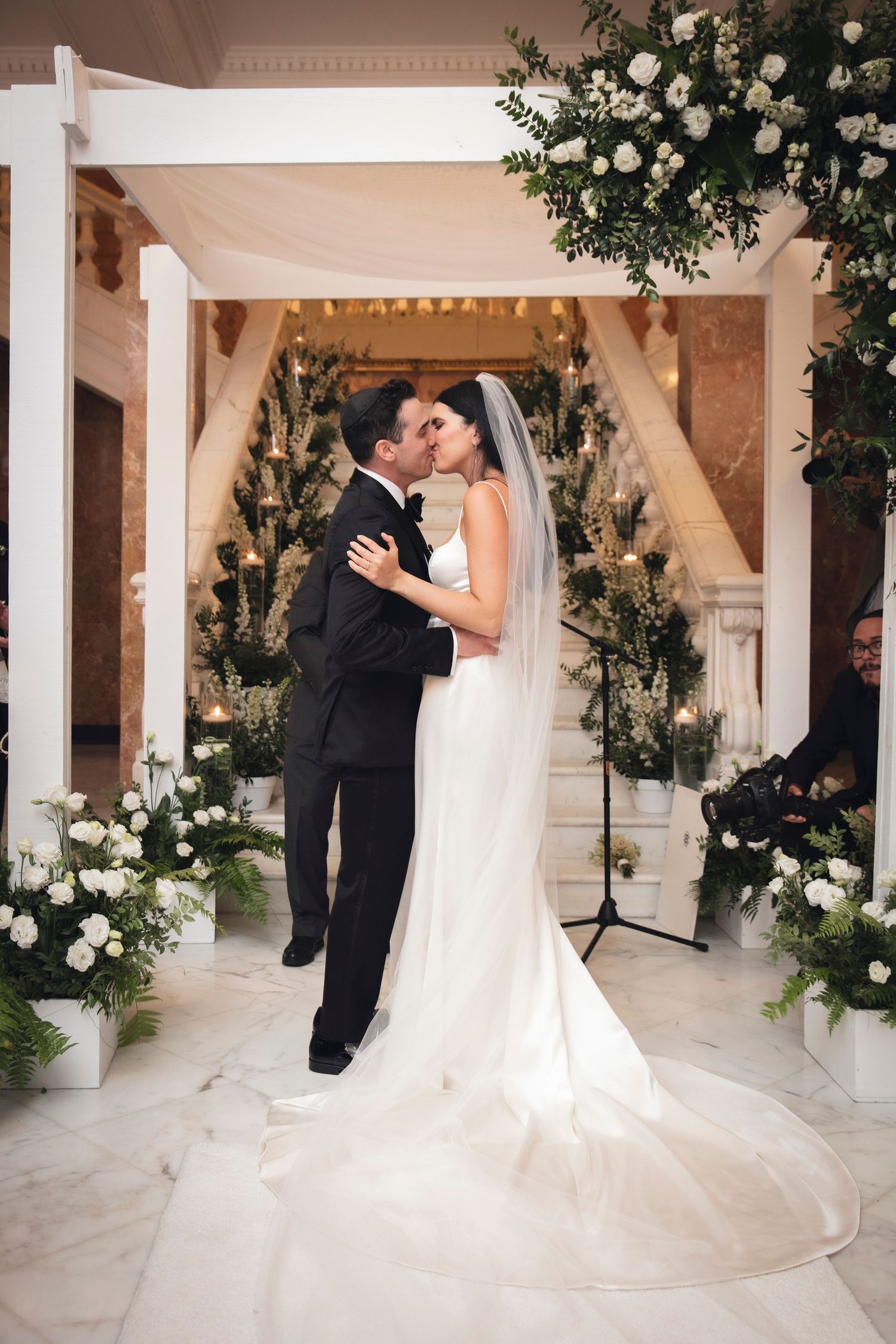 Wedding couple shares romantic moments under white columns adorned with elegant floral arrangements.