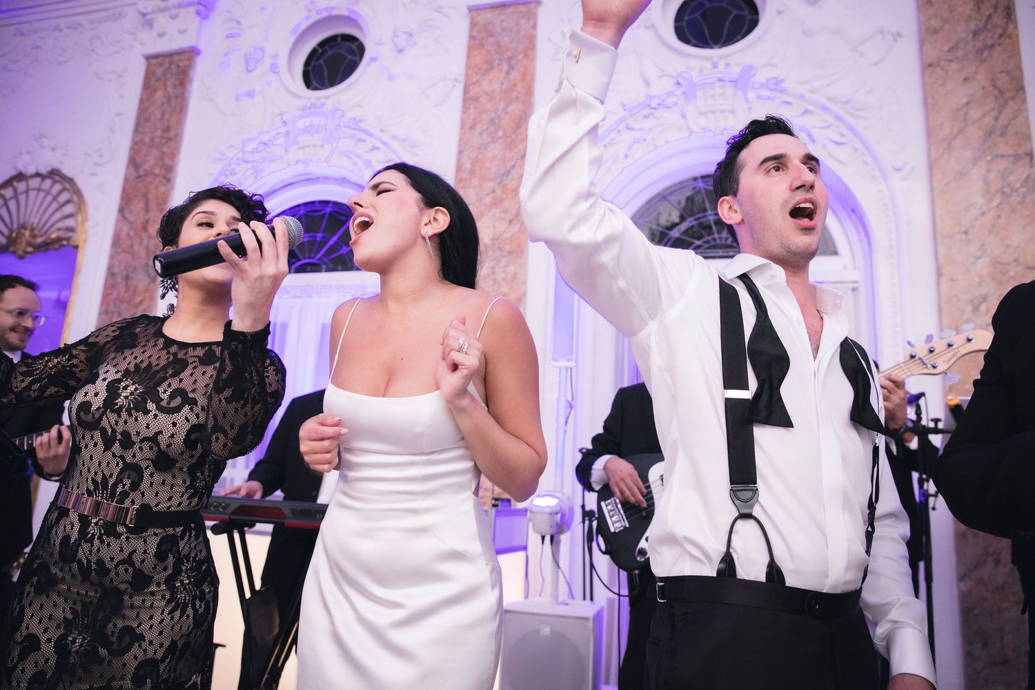 People dancing and laughing at an elegant wedding reception with purple uplighting and white columns.